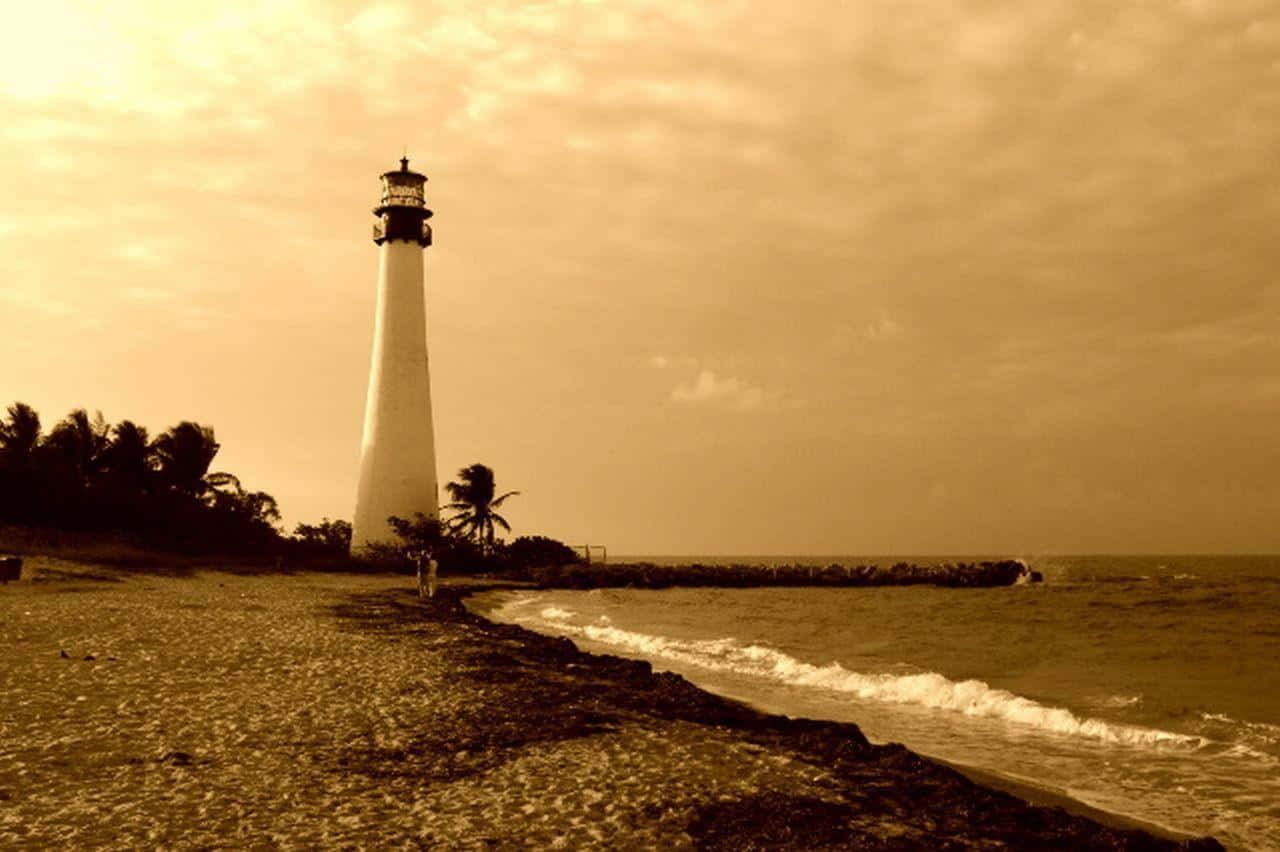 Sepia 桌布 壁紙 — 一座高大的白色燈塔矗立在岩石海灘上，夕陽西下，海浪在附近拍打，棕櫚樹在溫暖的棕褐色光線中形成剪影。