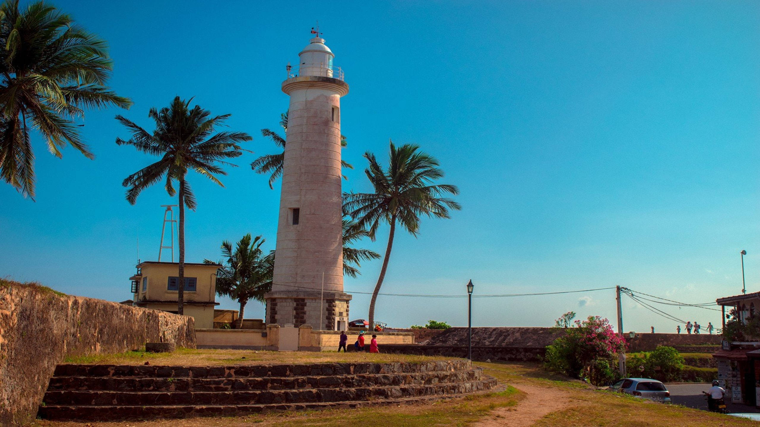 Sri Lanka 桌布 壁紙 — 一座高大的白色燈塔自豪地矗立在搖曳的棕櫚樹之間，在晴朗的藍天下，附近有石牆和遊客。