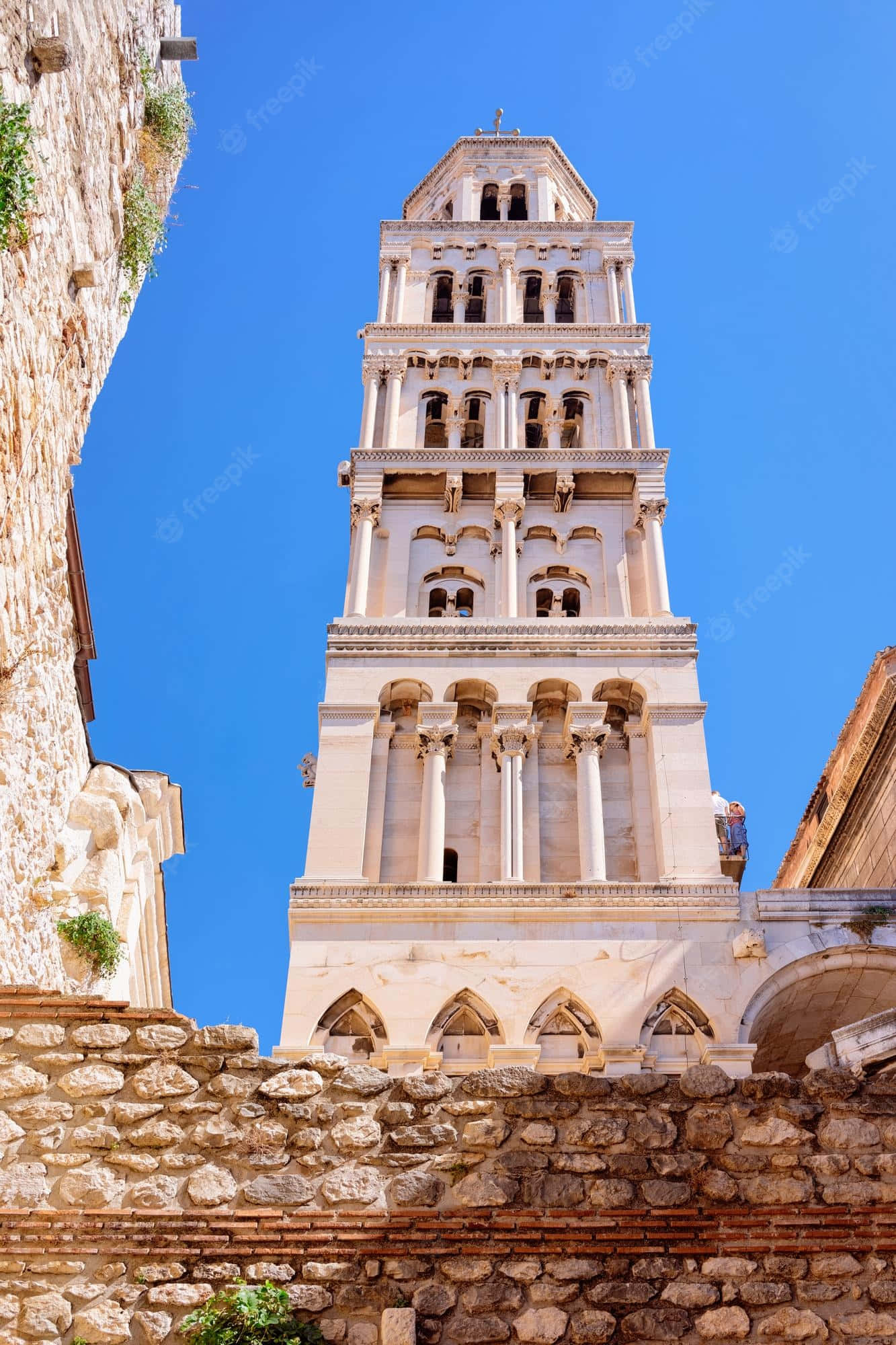 Croatia 桌布 壁紙 — 一座高大的白色石造鐘樓，在清澈的藍天下聳立，四周環繞著古老的石牆和拱門。