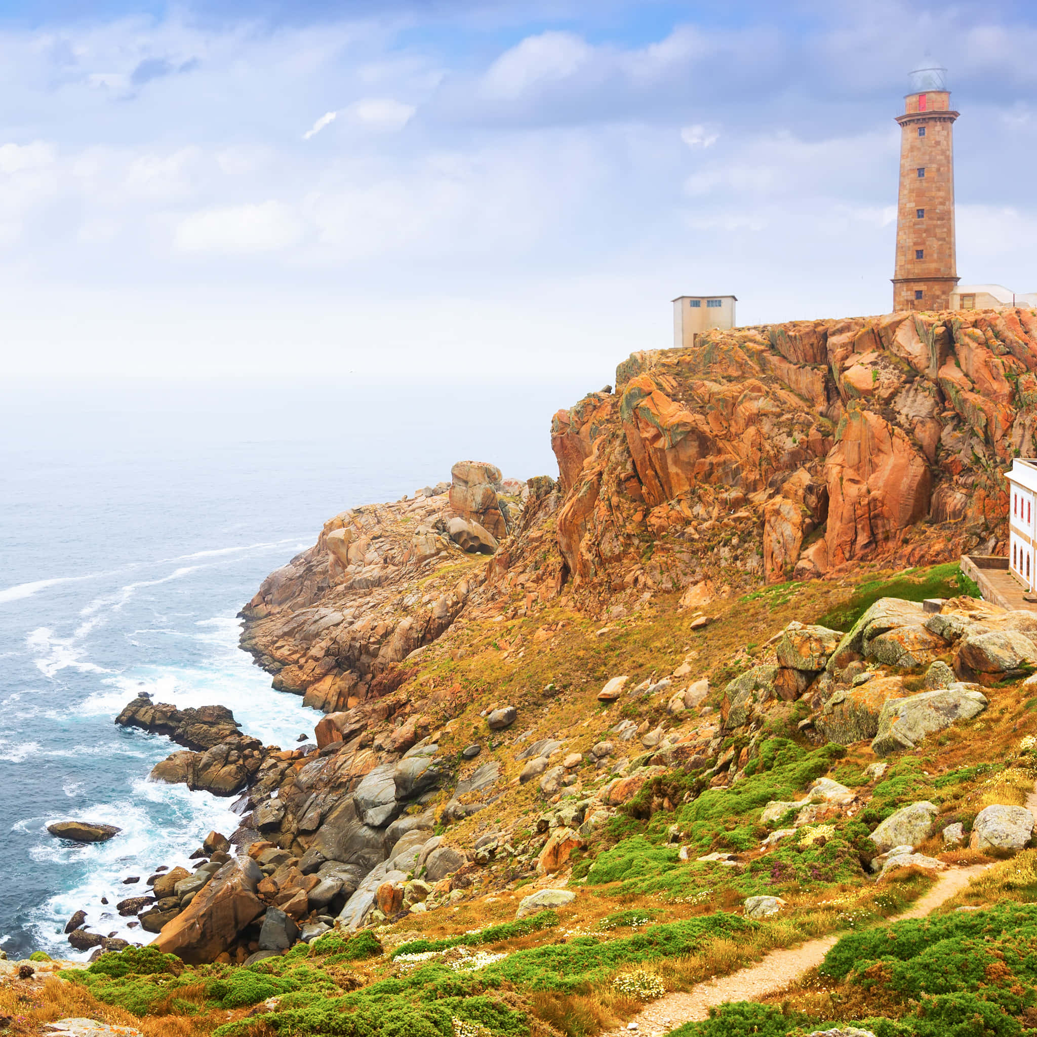 Lighthouse 桌布 壁紙 — 一座高大的石燈塔矗立在崎嶇的橙色懸崖上，俯瞰著部分多雲天空下翻騰的藍色大海。