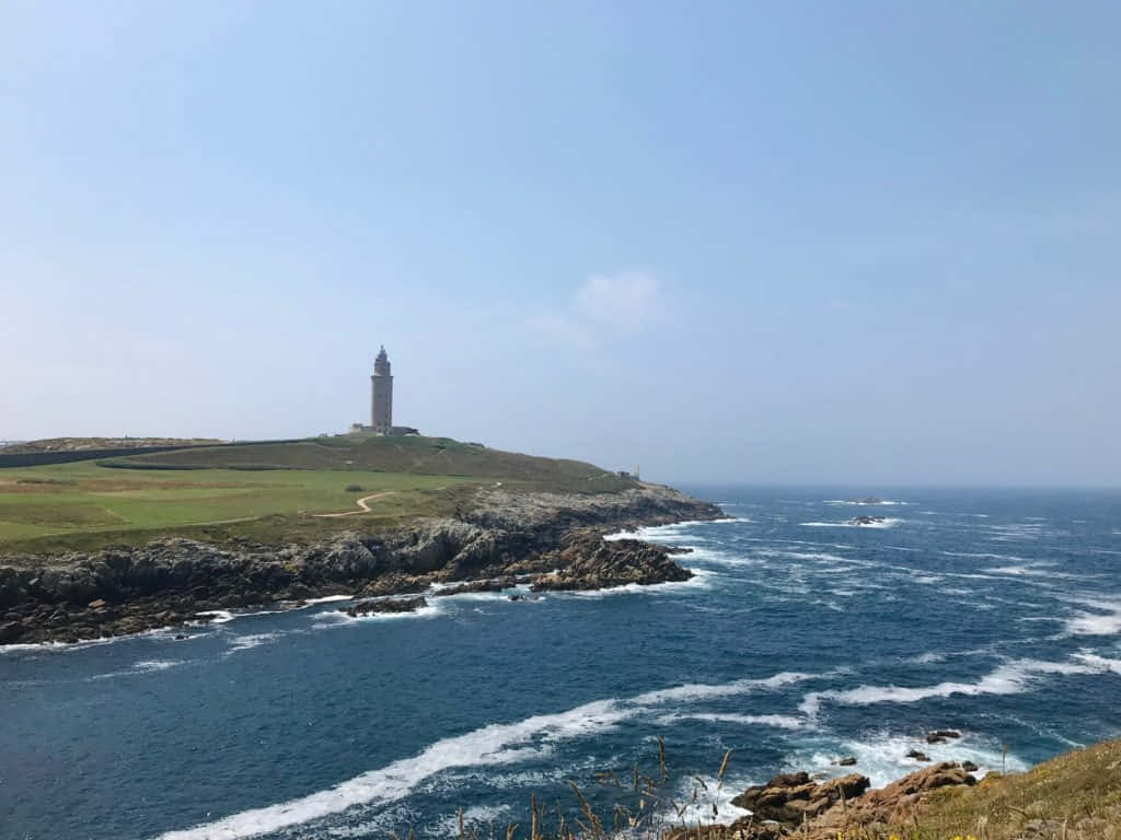 海景 桌布 壁紙 — 一座高大的石燈塔矗立在長滿青草的懸崖上，俯瞰著晴朗天空下拍打岩石海岸的洶湧藍色海浪。