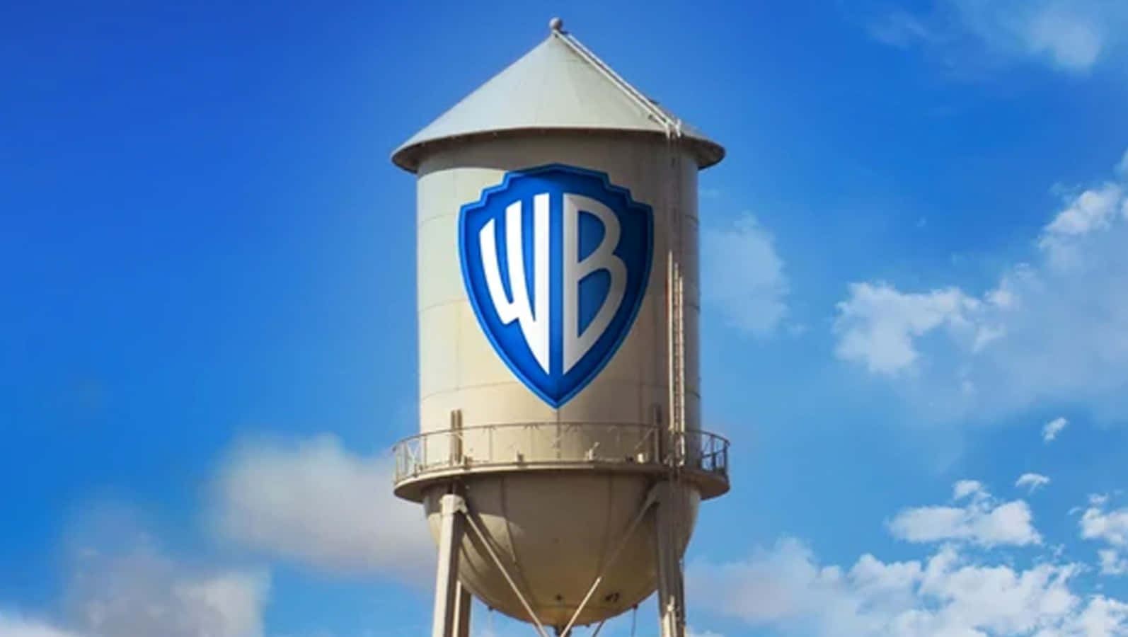 Warner Bros 桌布 壁紙 — 一座高大的米色水塔，上面有藍白相間的華納兄弟盾牌標誌，矗立在晴朗的藍天下，點綴著幾朵白雲。