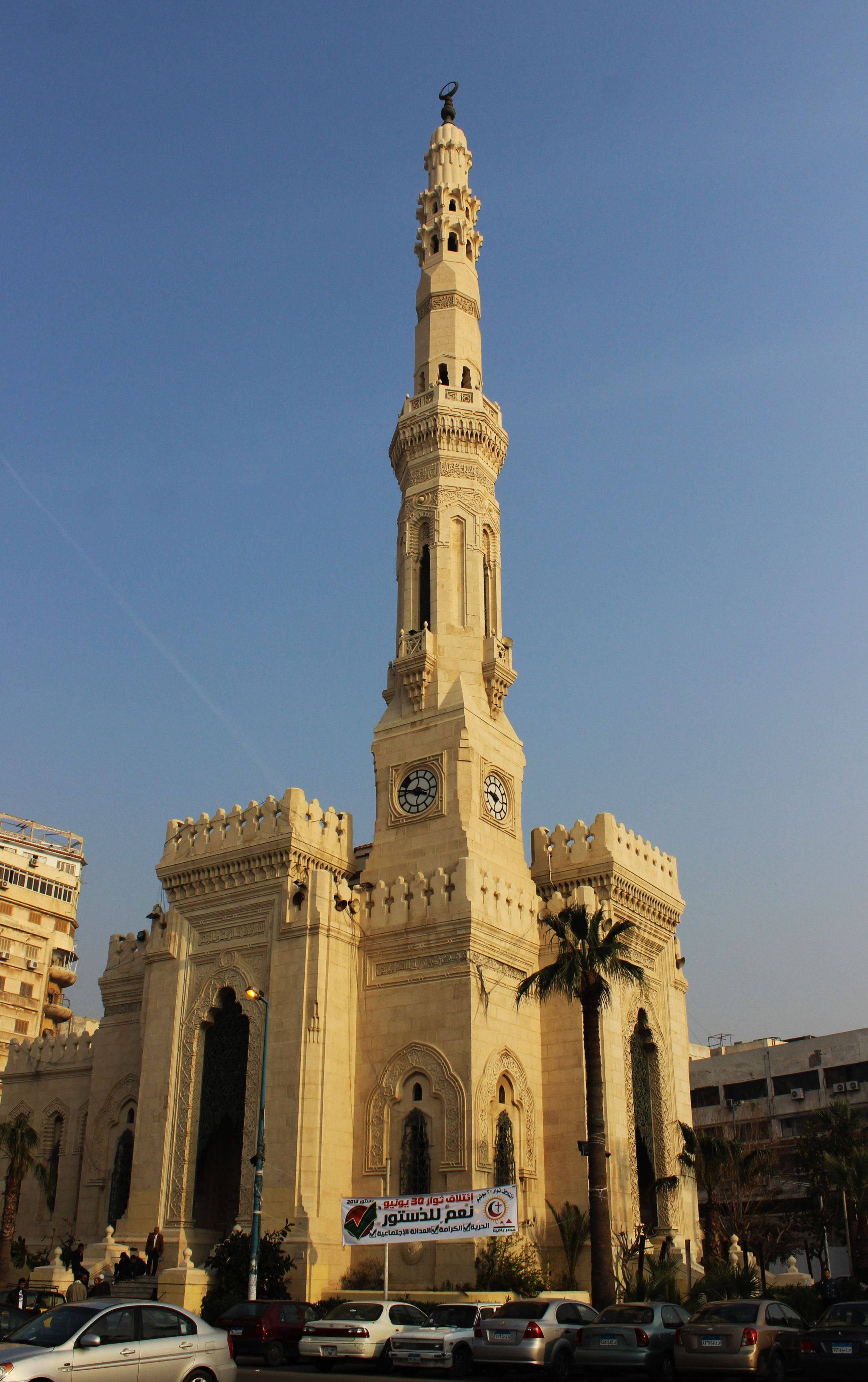 Alexandria 桌布 壁紙 — 一座高大的米色清真寺，帶有鐘樓和宣禮塔，矗立在晴朗的藍天下，下方有停放的汽車和棕櫚樹。
