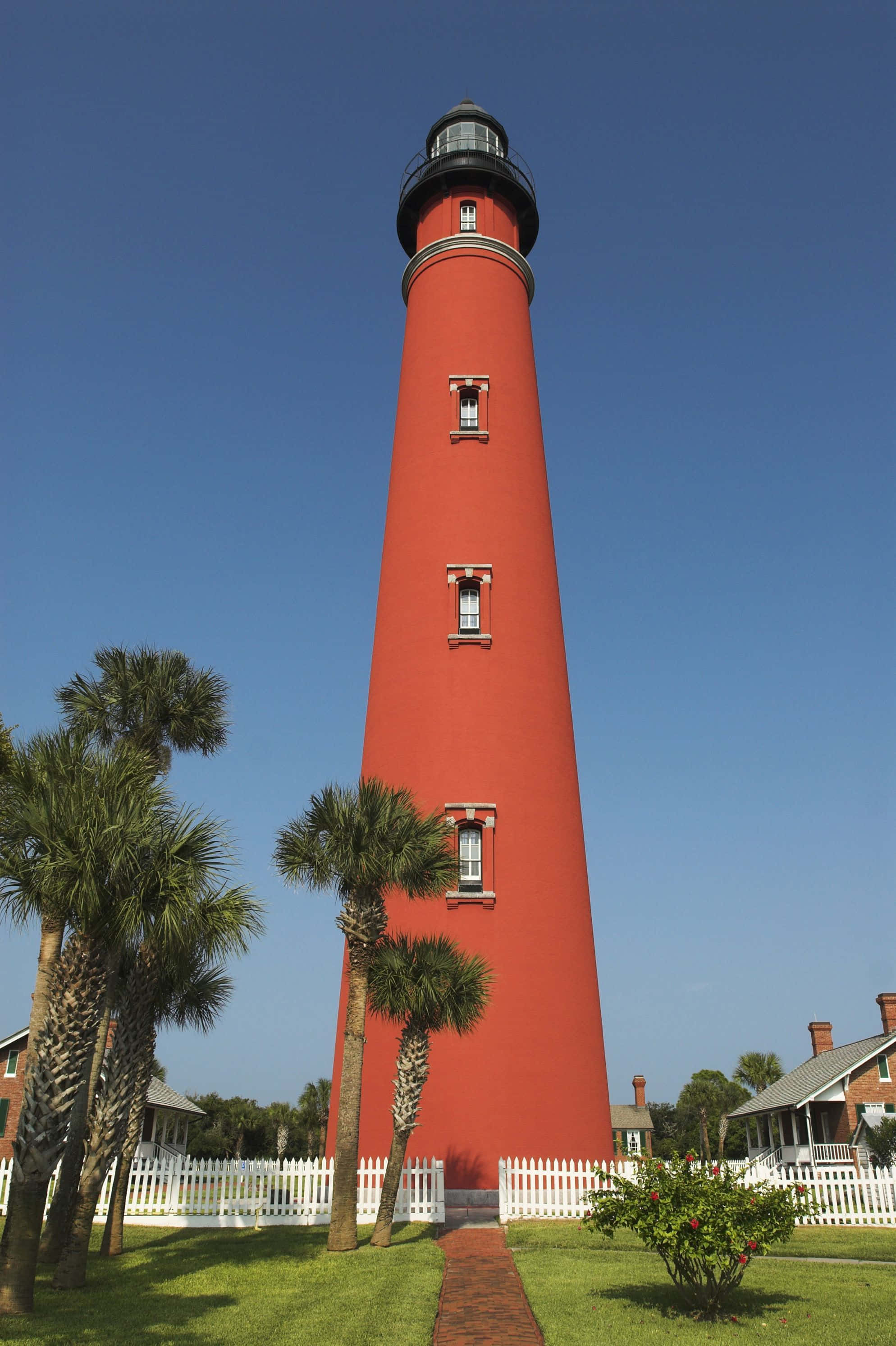 Lighthouse 桌布 壁紙 — 一座高大的紅色燈塔矗立在晴朗的藍天下，周圍環繞著棕櫚樹和白色尖樁籬笆。