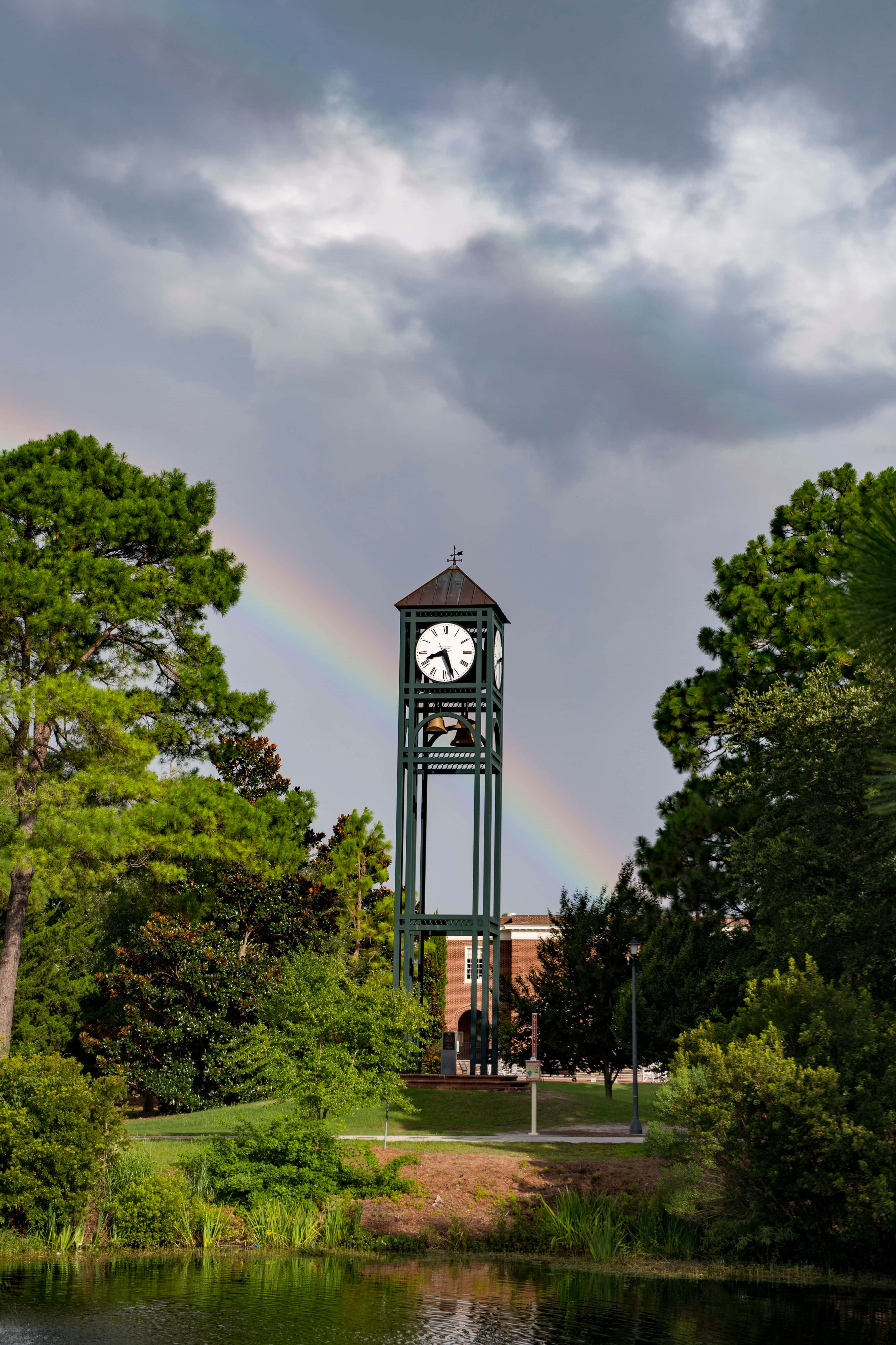 Clock 桌布 壁紙 — 一座高大的綠色鐘樓矗立在茂密的公園中，背後有鮮豔的彩虹在陰雲密佈的天空下拱起。