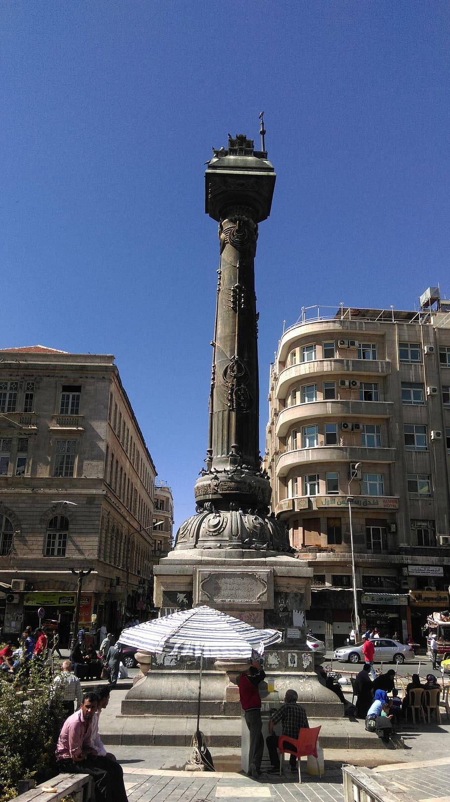 Syria 桌布 壁紙 — 一座高大的華麗柱式紀念碑矗立在繁忙的城市廣場上，天空湛藍，人們在附近休憩。
