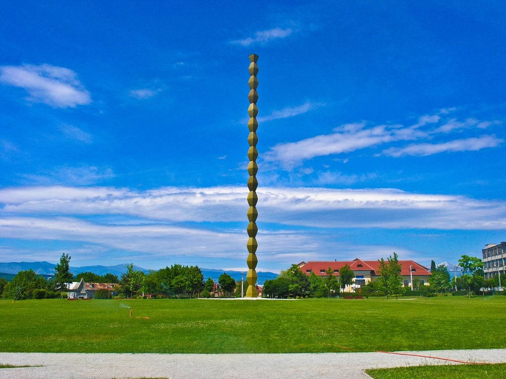 Romania 桌布 壁紙 — 一座高大的金色螺旋雕塑矗立在充滿活力的綠色公園中，天空湛藍，白雲點點。
