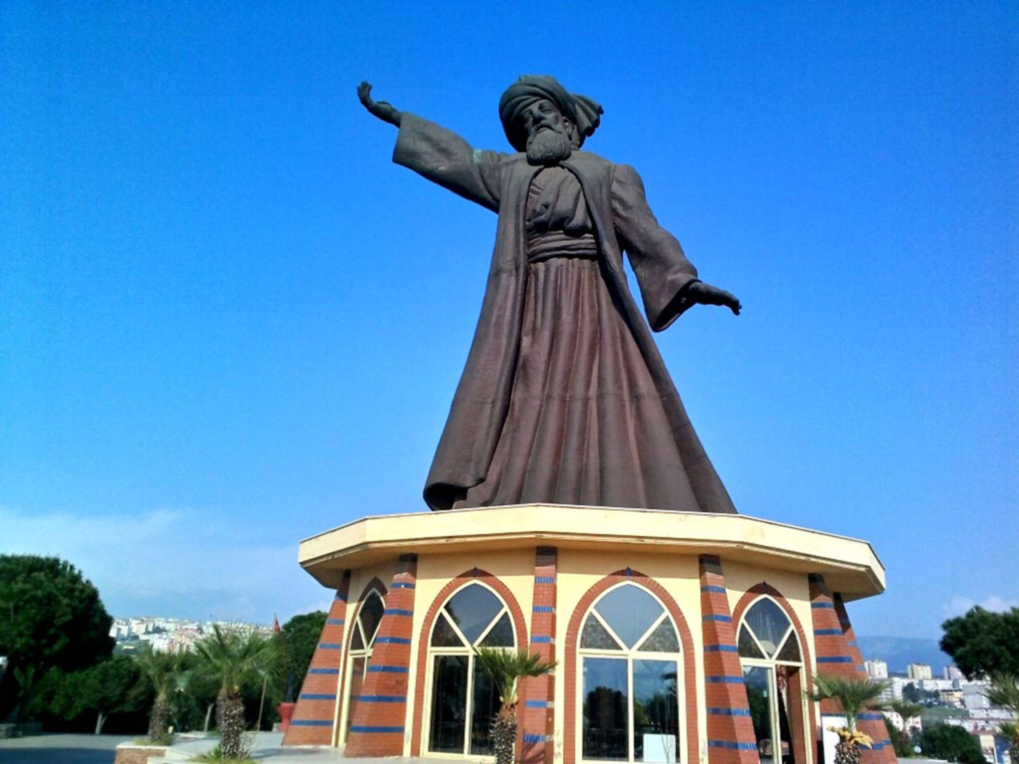 Konya 桌布 壁紙 — 一座高大的鬍鬚男子青銅雕像，身穿傳統長袍，站在色彩繽紛的瓷磚基座上，背景是清澈的藍天。