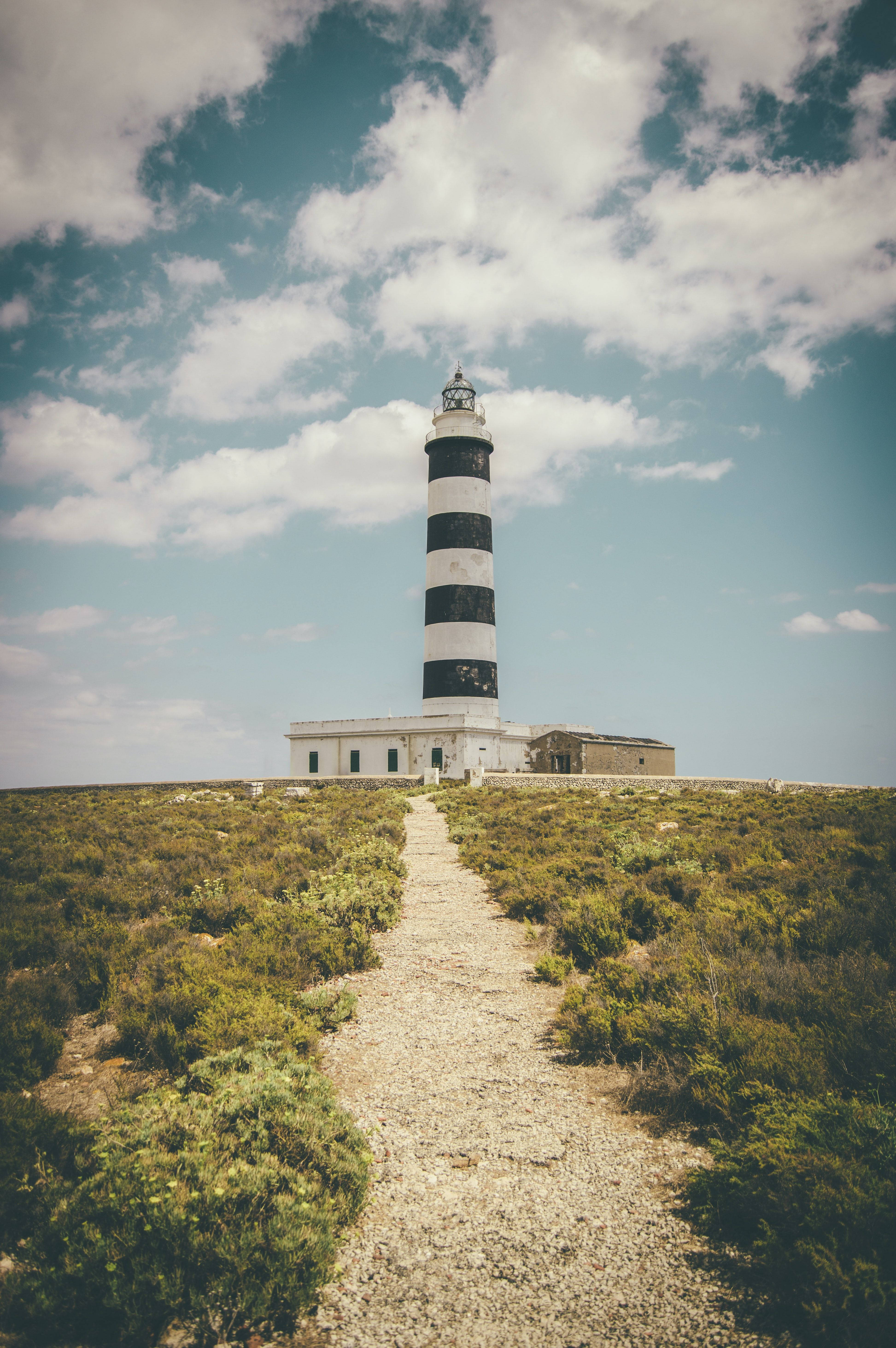 Lighthouse 桌布 壁紙 — 一座高大的黑白條紋燈塔矗立在長滿草的懸崖上，天空多雲，有一條碎石路通往燈塔。
