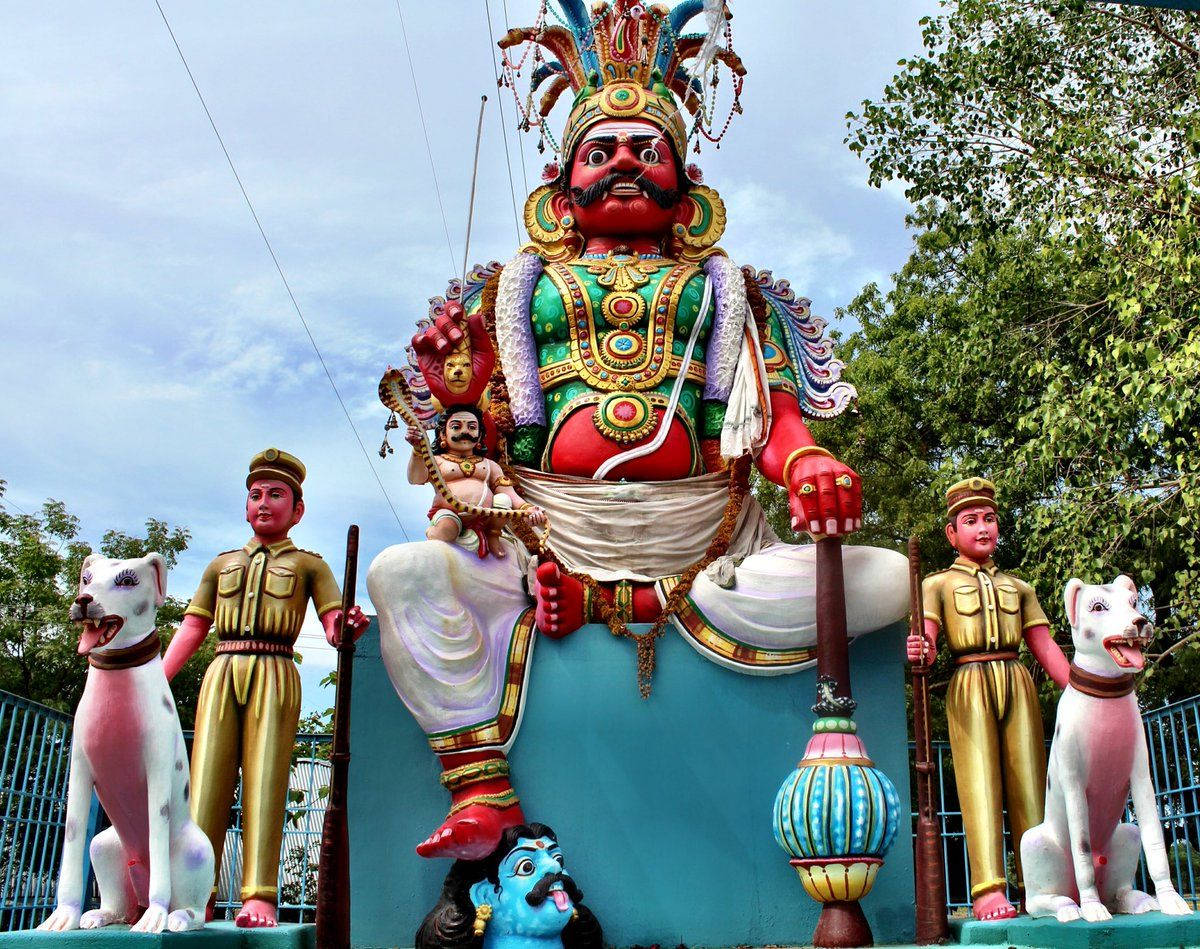 statue 桌布 壁紙 — 一座高聳、色彩鮮豔的印度教神像，紅臉戴華麗皇冠，兩旁有兩名士兵和白色狗，天空部分多雲。