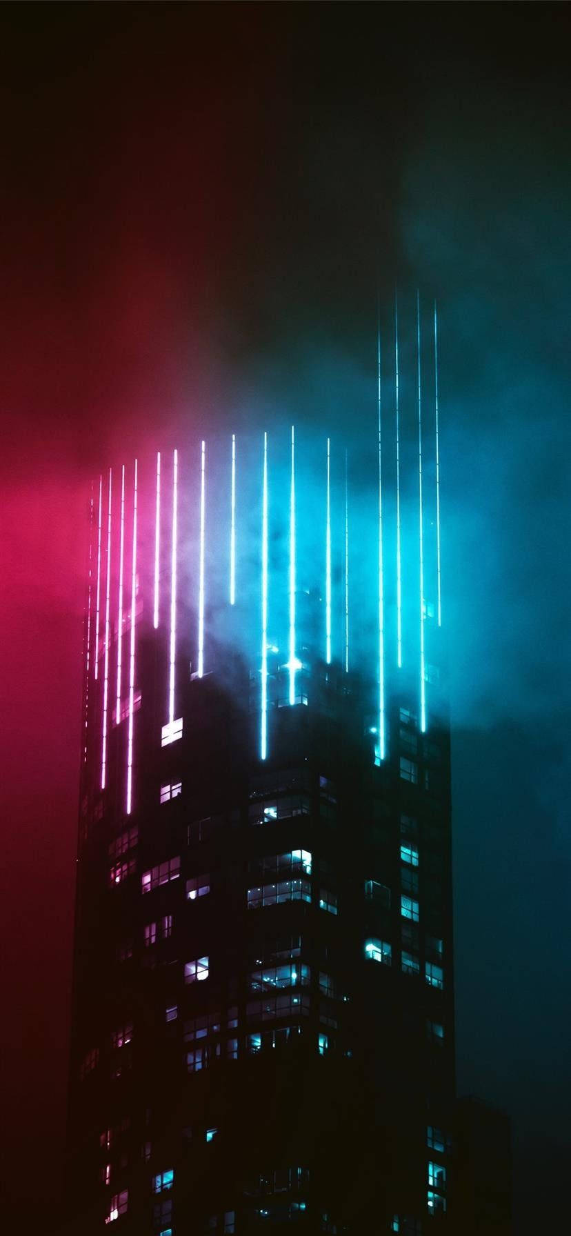 lights 桌布 壁紙 — 一座高聳入雲的摩天大樓在夜晚被粉紅色和青色的垂直霓虹燈光瀑布照亮，背景是煙霧瀰漫的天空。