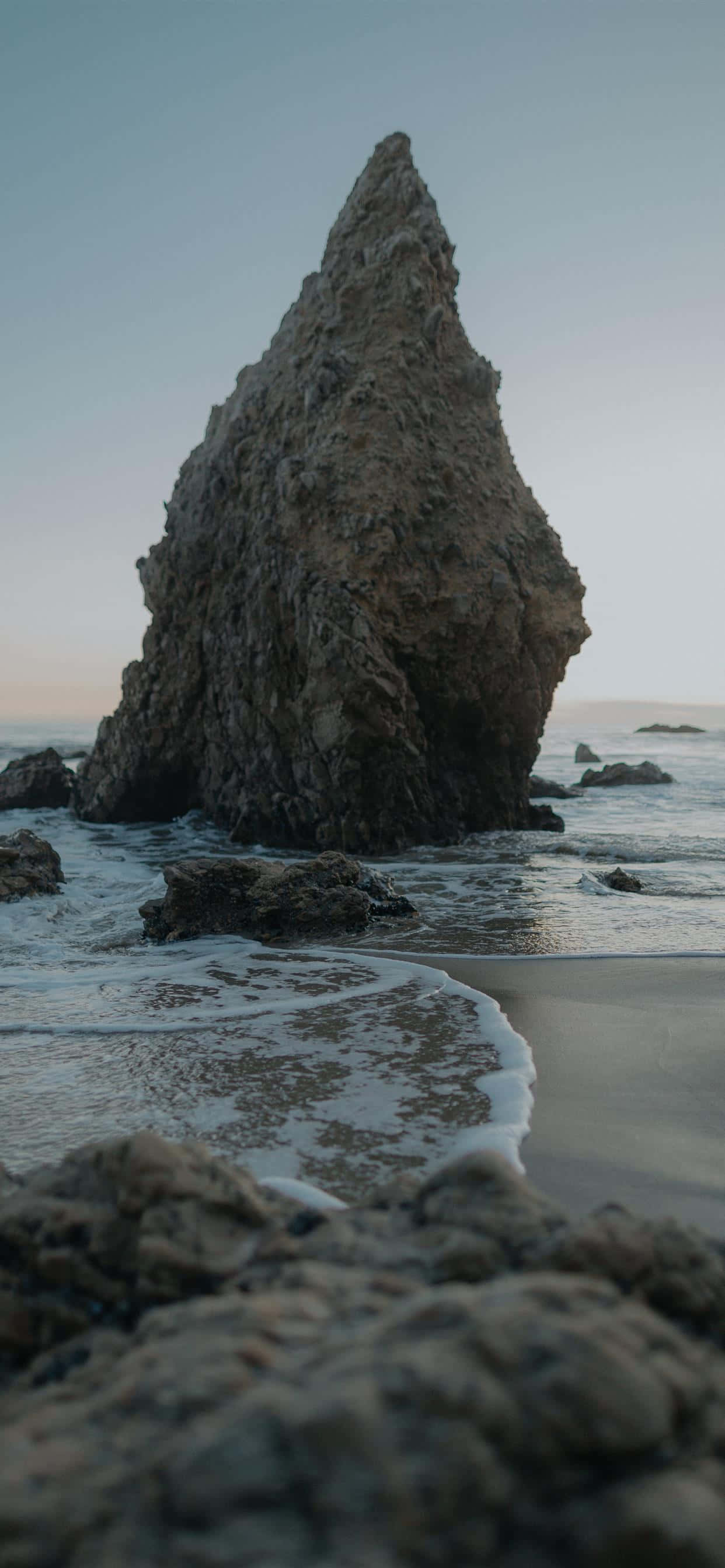 Malibu California 桌布 壁紙 — 一座高聳崎嶇的岩石形成物孤獨地矗立在霧氣瀰漫的海灘上，溫柔的海浪在黃昏時分拍打著它的底部。