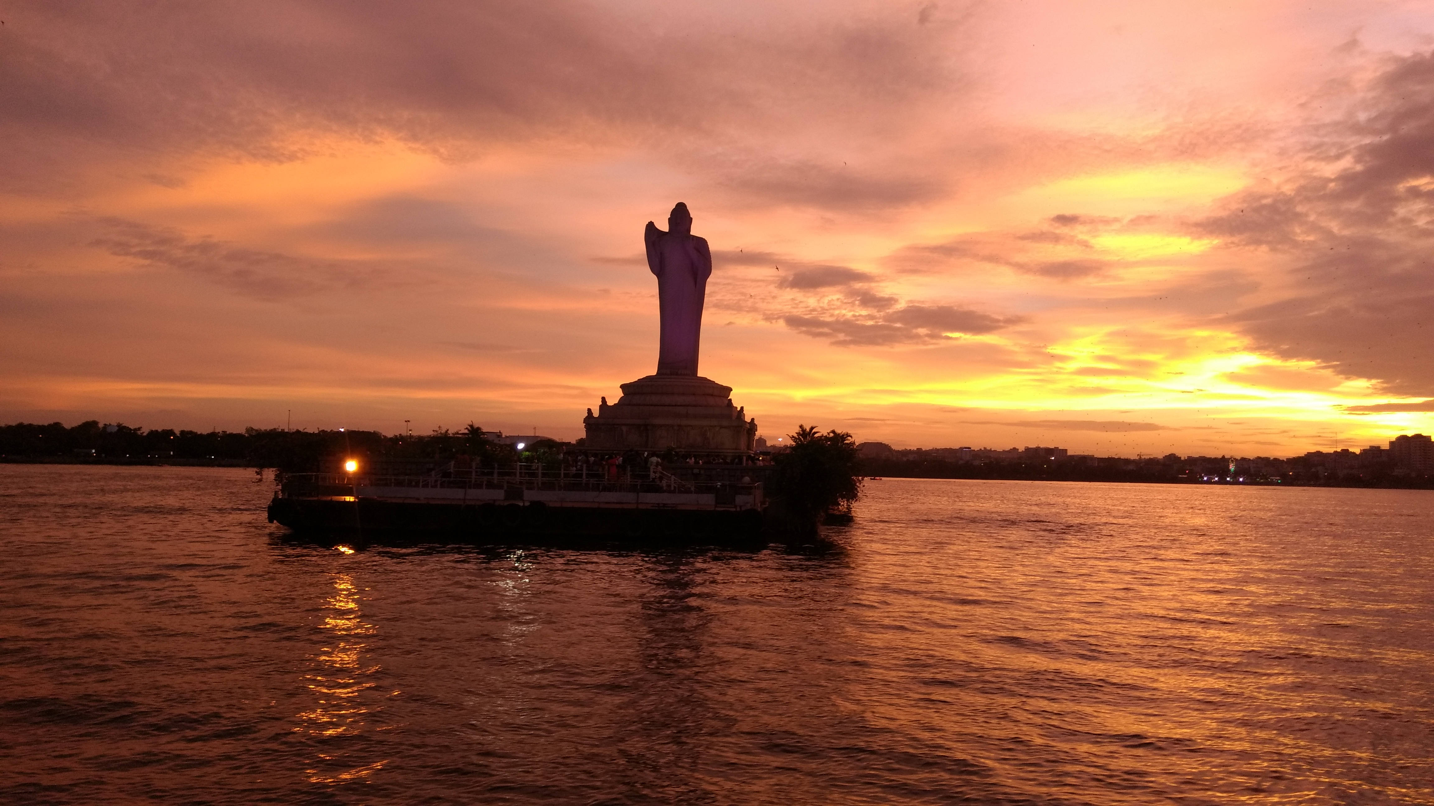 Hyderabad 桌布 壁紙 — 一座高聳的佛像在日落時分靜靜地矗立在小島上，金色的光芒灑在平靜的水面上。