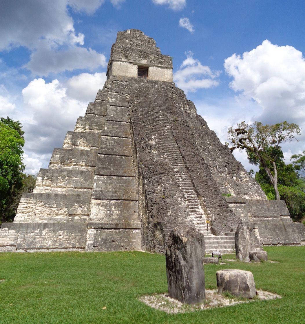 Guatemala 桌布 壁紙 — 一座高聳的古老瑪雅金字塔在明亮的藍天白雲下拔地而起，周圍環繞著茂密的綠草和樹木。
