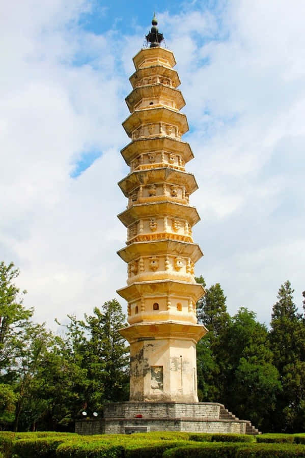 Building 桌布 壁紙 — 一座高聳的古老石塔，層層疊疊的屋簷，在部分多雲的天空下，被茂密的綠樹環繞，優雅地矗立著。