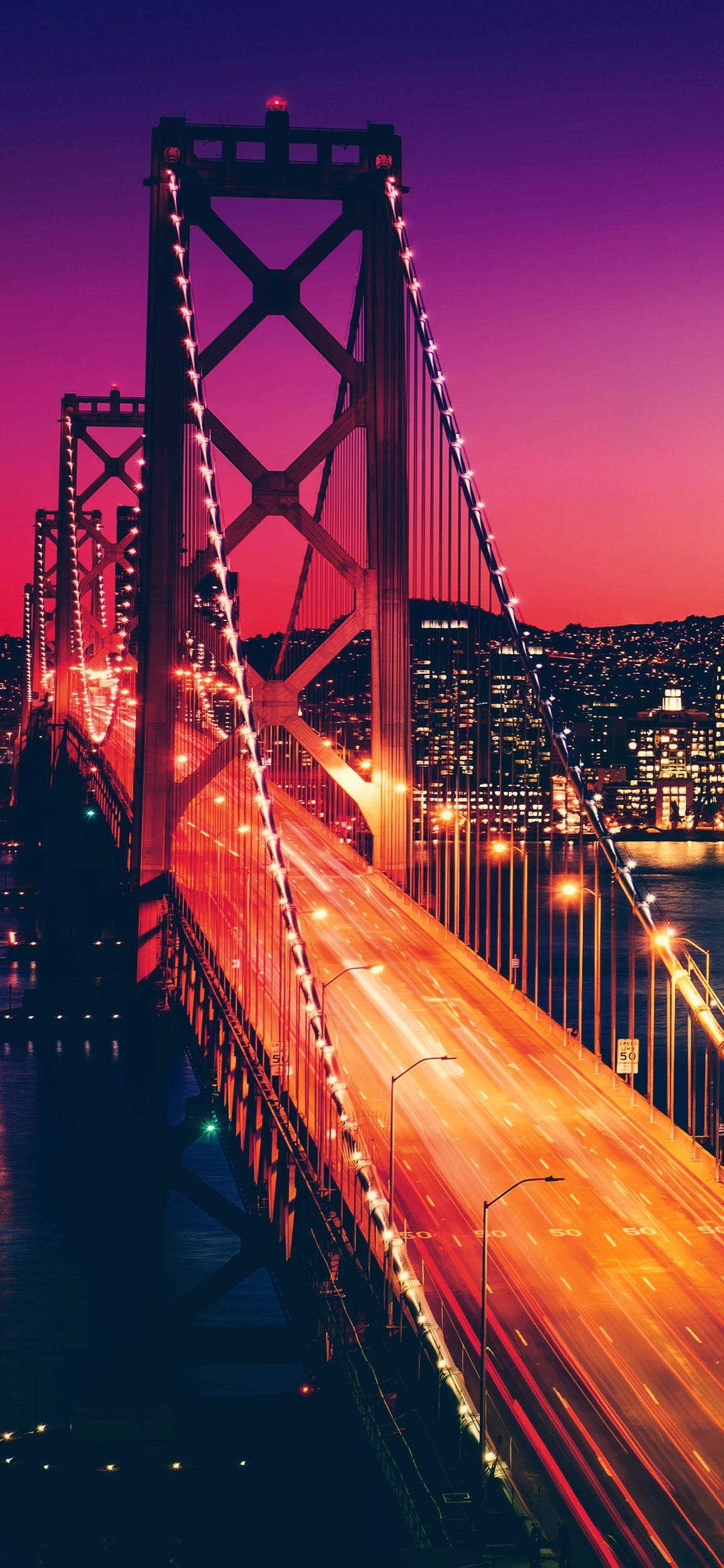 San Francisco Phone 桌布 壁紙 — 一座高聳的吊橋在充滿活力的紫色和粉紅色暮色天空中閃爍著橙色交通燈的條紋。