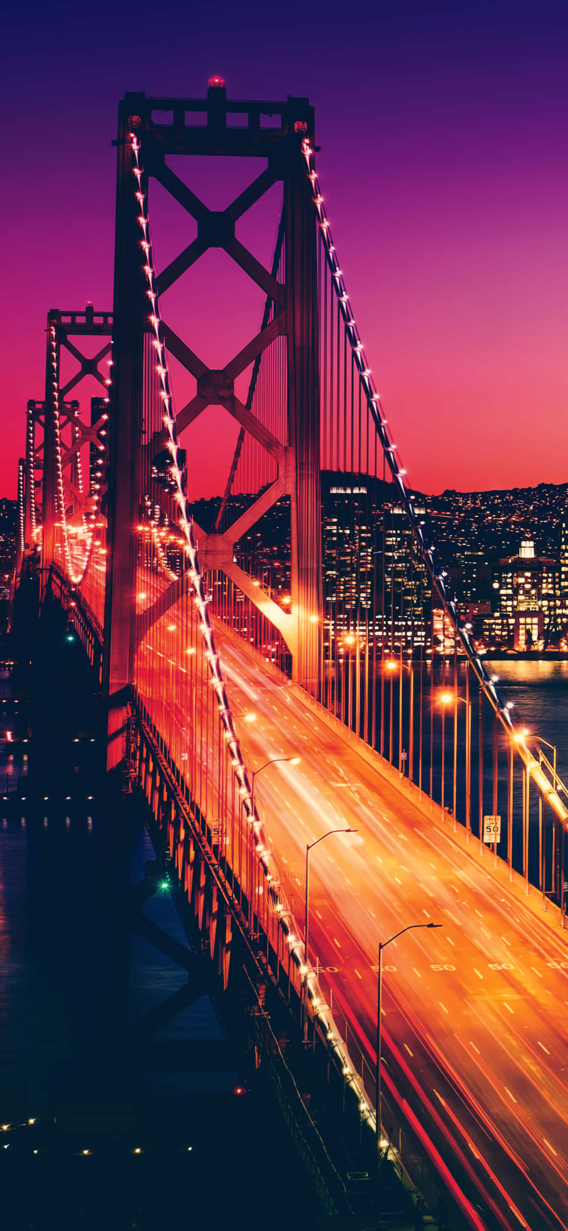 iPhone X California 桌布 壁紙 — 一座高聳的吊橋在充滿活力的紫色和粉紅色暮色天空中閃爍著橙色交通燈的條紋。