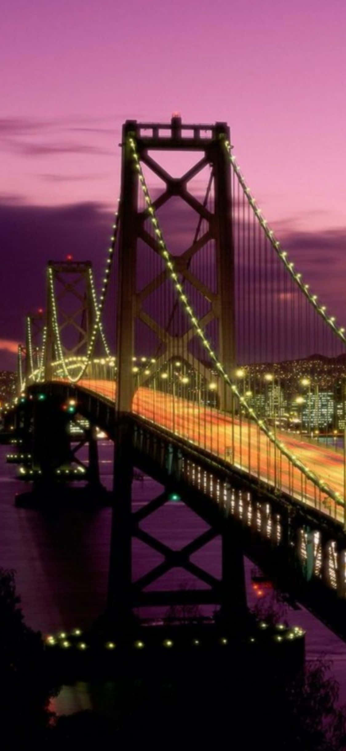 iPhone Xs California 桌布 壁紙 — 一座高聳的吊橋在充滿活力的紫色暮色天空下，散發著溫暖的光芒，映照著城市景觀。