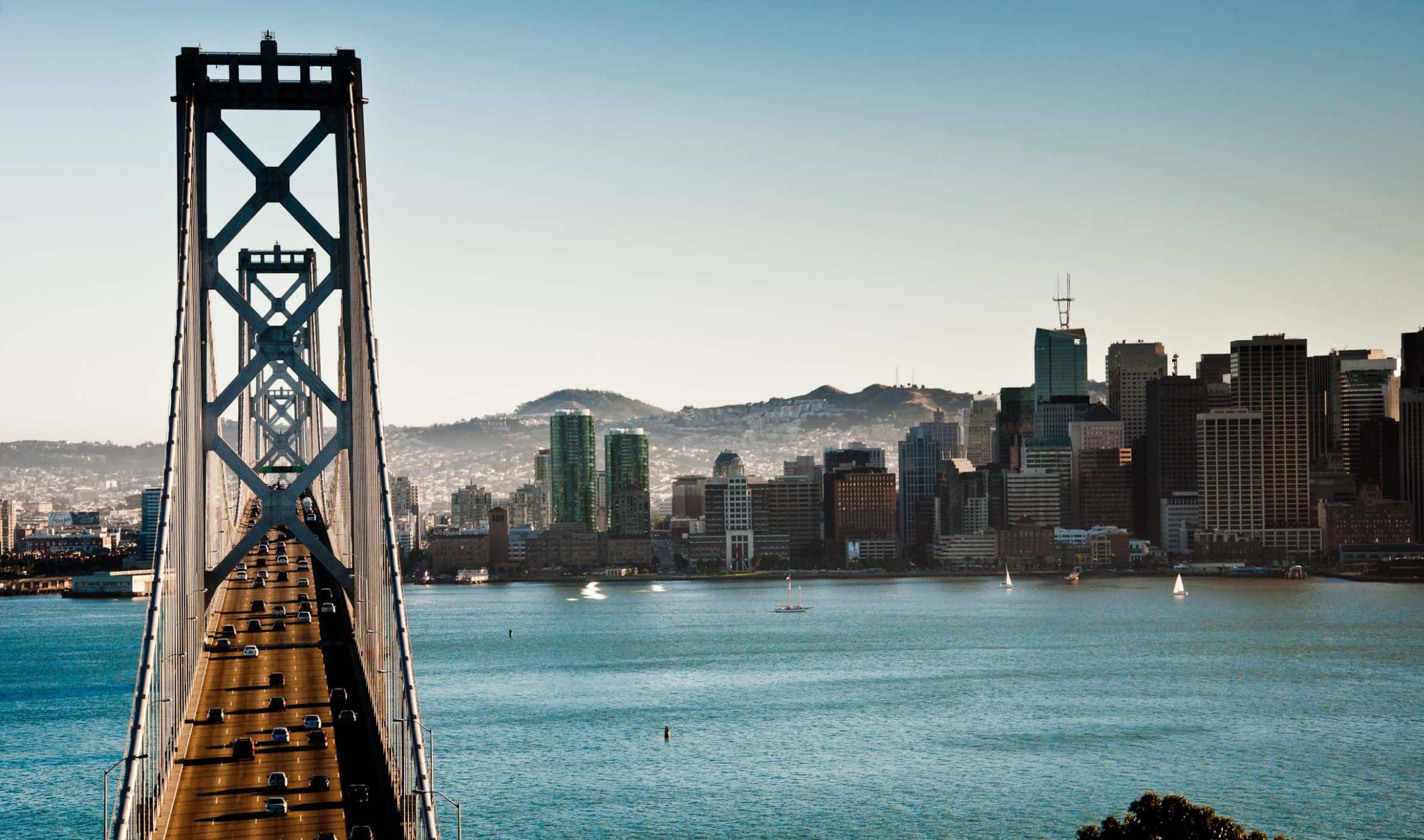 2440x1440 San Francisco 桌布 壁紙 — 一座高聳的吊橋橫跨在碧綠色的水面上，通往陽光明媚的城市天際線，帆船在橋下滑行。