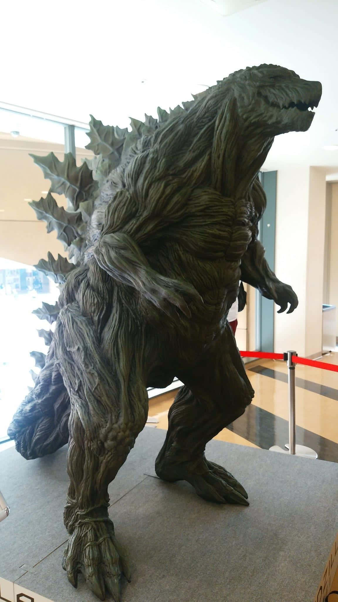 Godzilla Earth 桌布 壁紙 — 一座高聳的哥吉拉雕像，帶有紋理的綠褐色皮膚和尖刺背部，在現代室內大廳展出。