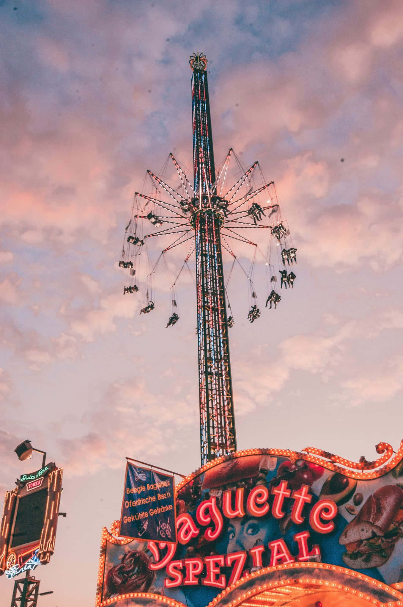 Carnival 桌布 壁紙 — 一座高聳的嘉年華遊樂設施，帶有搖擺座椅，在美食攤位招牌上方的柔和日落天空中閃耀。