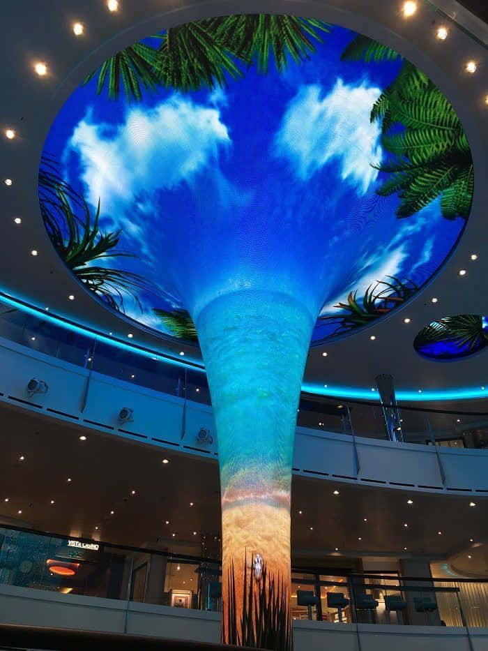 Carnival Vista 桌布 壁紙 — 一座高聳的室內瀑布展示了一個生動的熱帶天空，棕櫚葉和雲朵在沙灘場景之上。