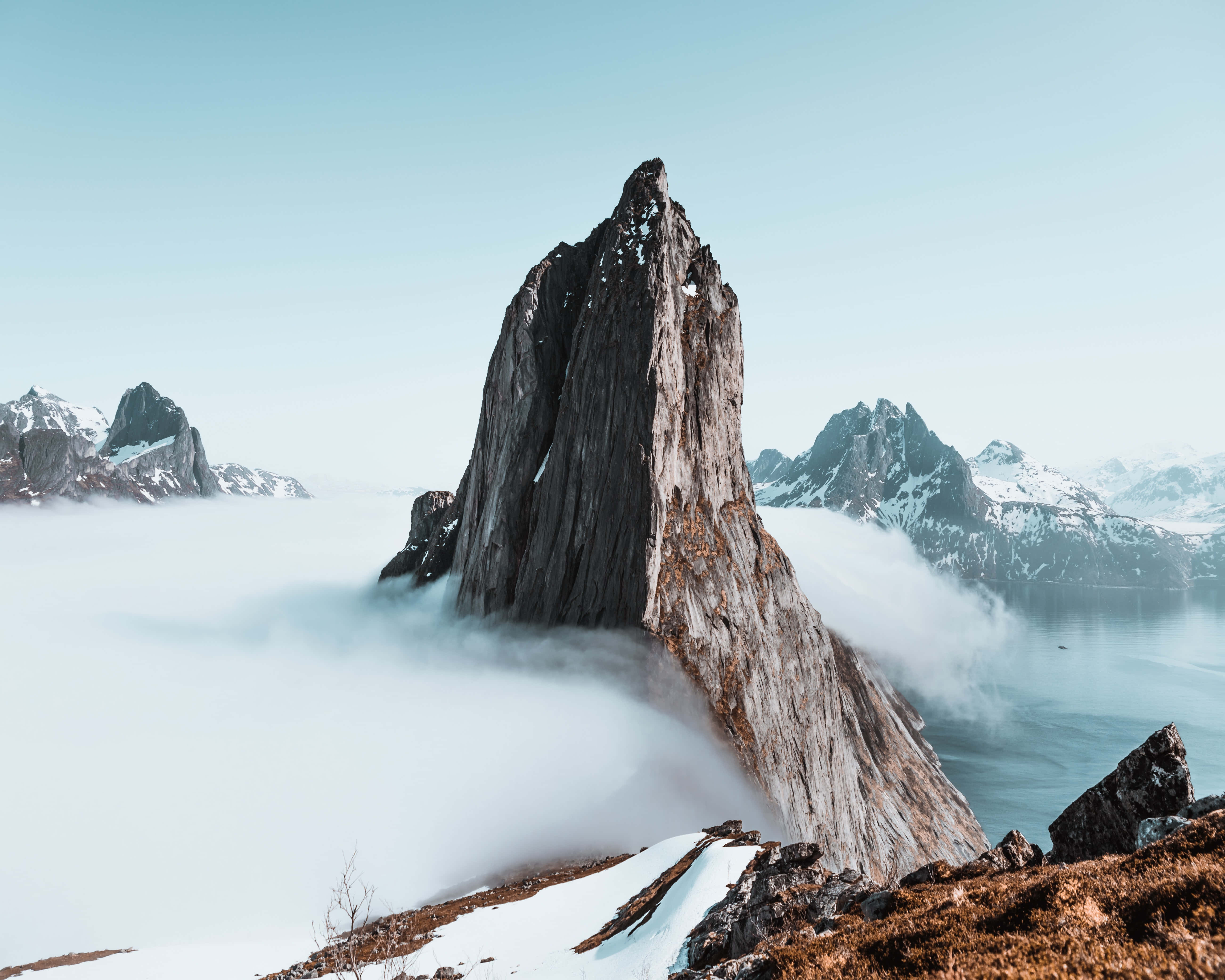 風景 桌布 壁紙 — 一座高聳的岩石山峰從雲海中戲劇性地升起，背景是白雪覆蓋的山脈和寧靜的水面。