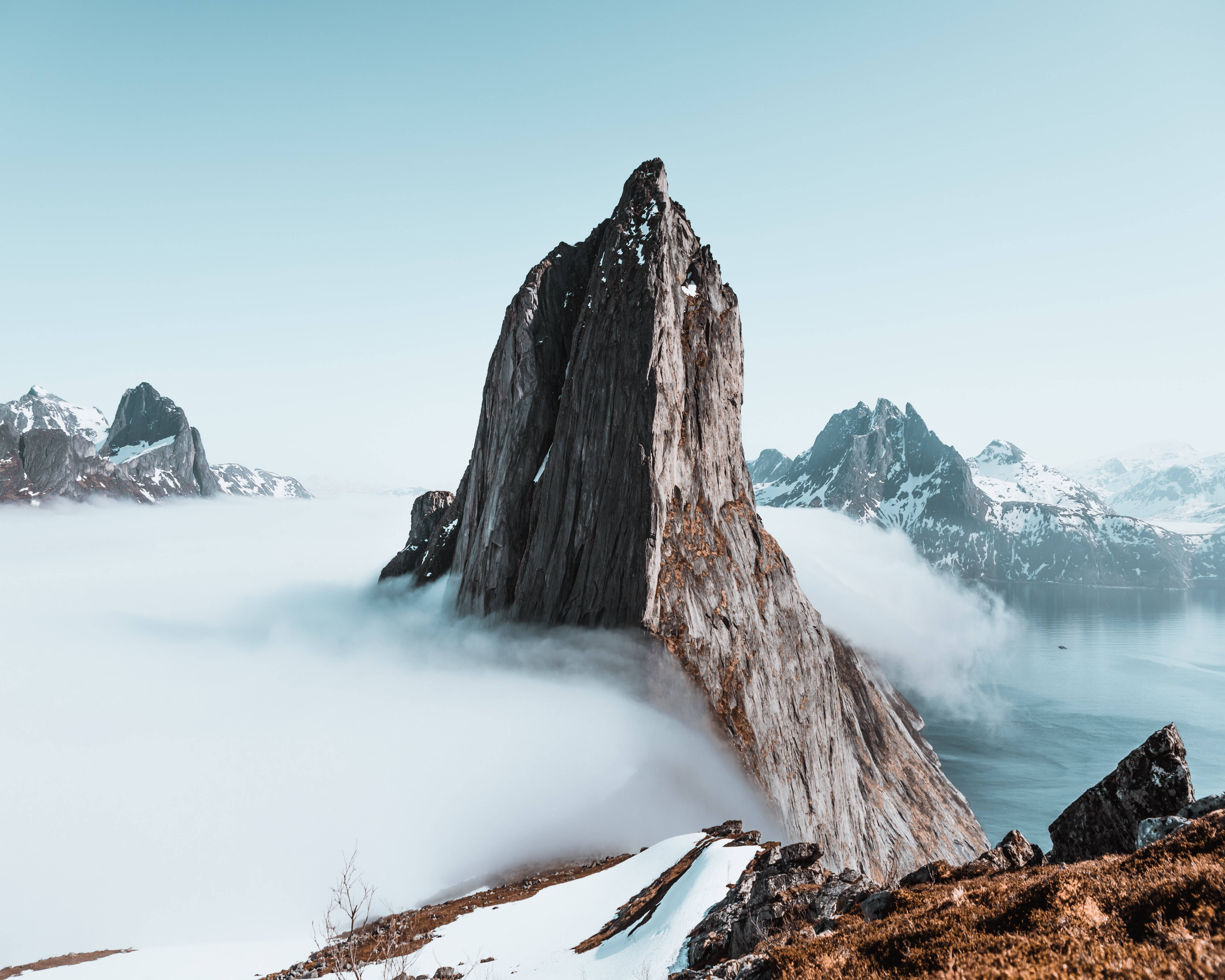 雲朵 桌布 壁紙 — 一座高聳的岩石山峰穿過雲海，遠處有積雪的山峰和寧靜的水面。