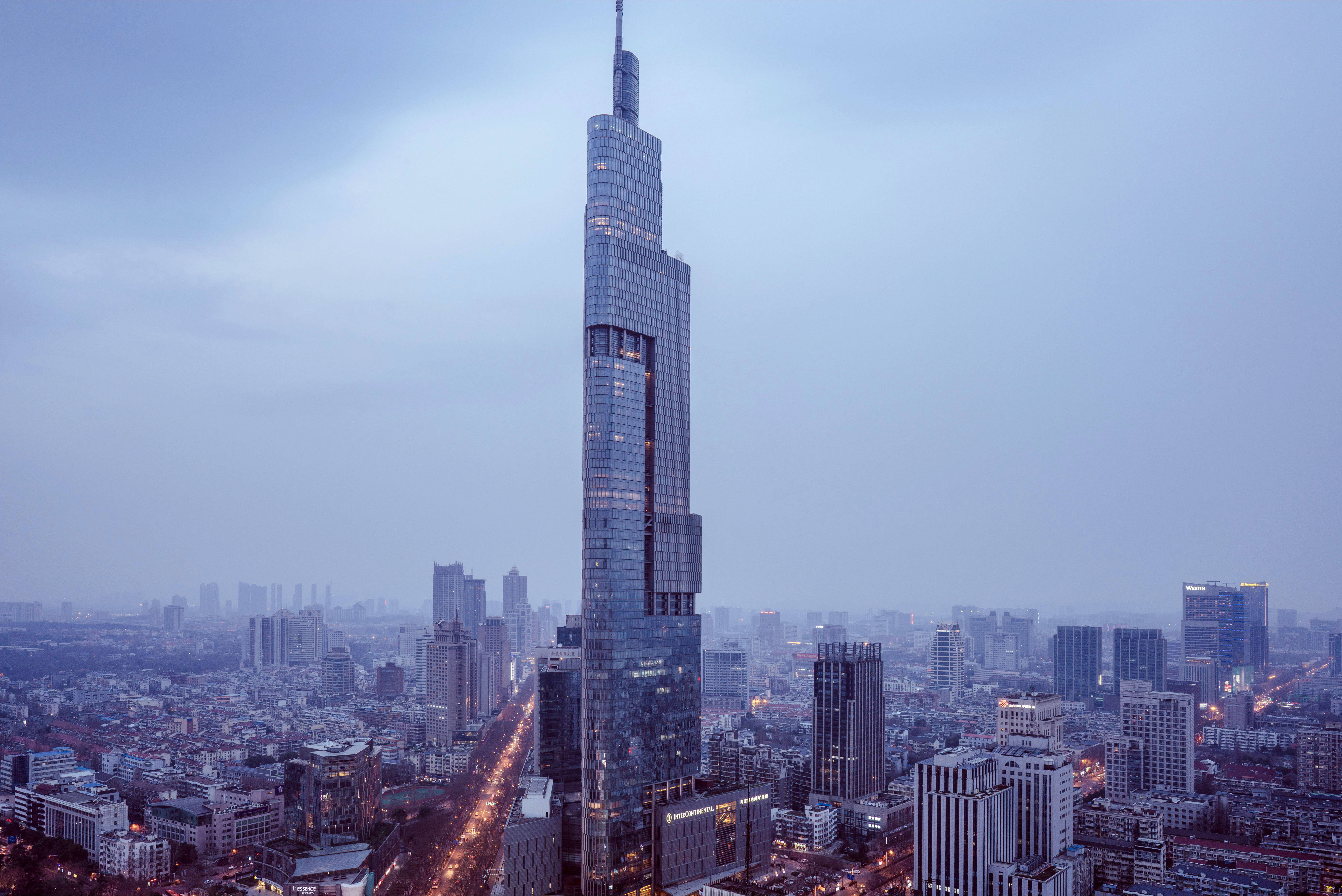 Nanjing 桌布 壁紙 — 一座高聳的摩天大樓主宰著廣闊的城市景觀，在朦朧的暮色天空下，下方有發光的街燈。