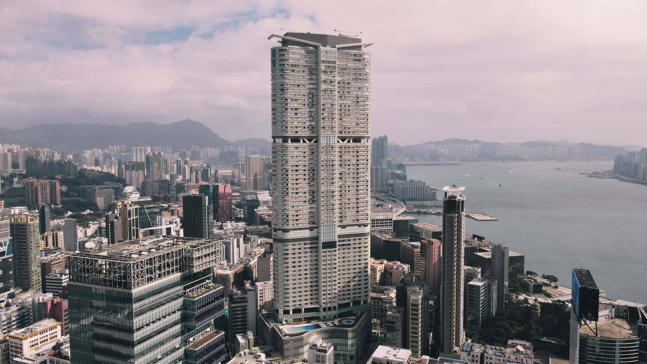 香港 桌布 壁紙 — 一座高聳的摩天大樓主導著密集的城市天際線，俯瞰著一個港口，遠處有山脈，天空多雲。