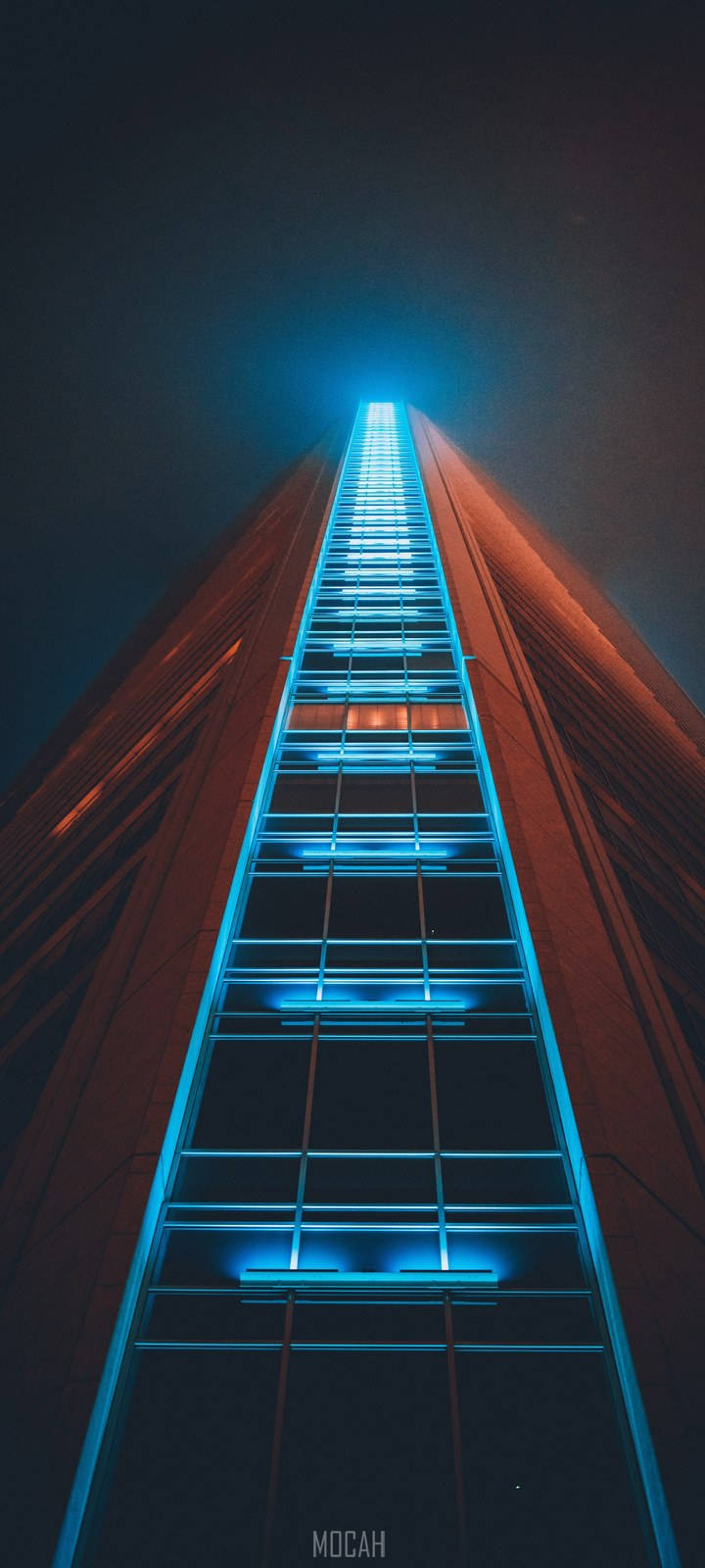 Skyscraper 桌布 壁紙 — 一座高聳的摩天大樓升入夜空，其玻璃外牆在黑暗的城市背景下閃耀著充滿活力的藍色燈光。