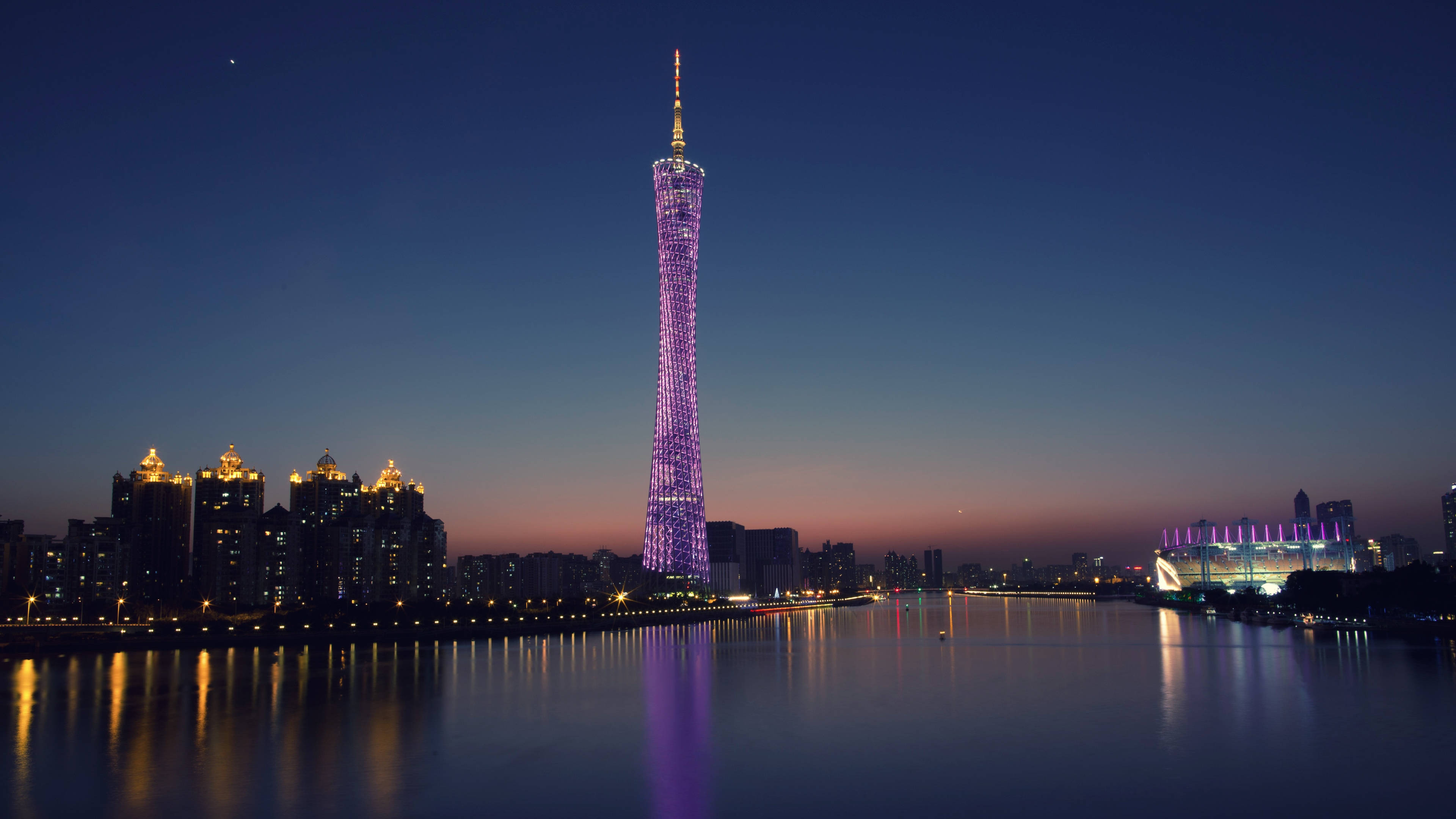 Guangzhou 桌布 壁紙 — 一座高聳的摩天大樓在暮色天空中散發紫色光芒，倒映在平靜的河水上，城市燈光在下方閃爍。