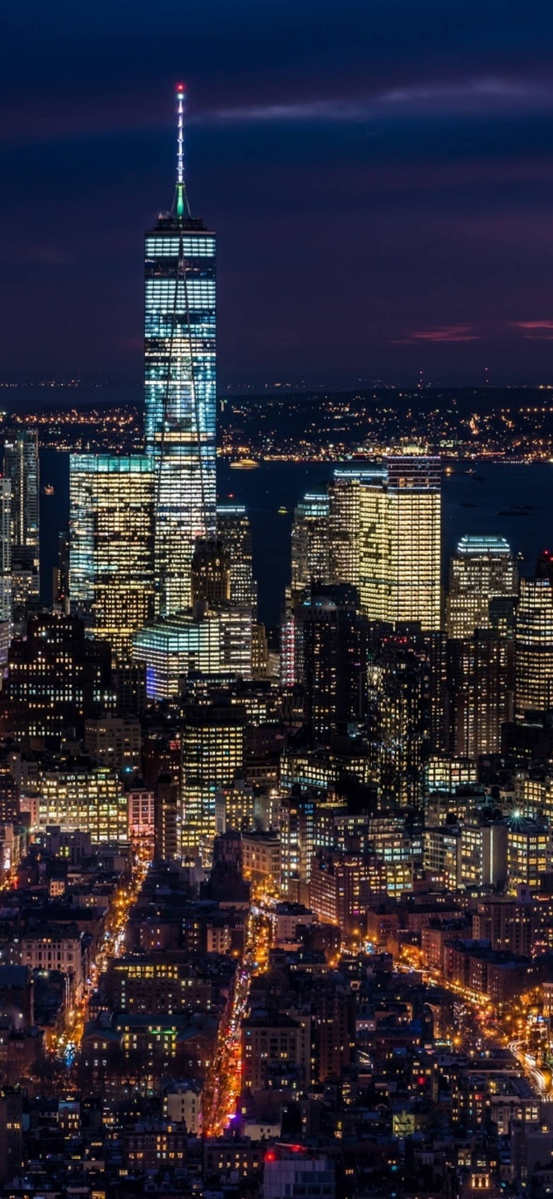 new york city iPhone x 桌布 壁紙 — 一座高聳的摩天大樓在深紫色的夜空下閃爍著藍光，下方是密集的城市景觀，建築物燈火通明，街道閃爍發光。