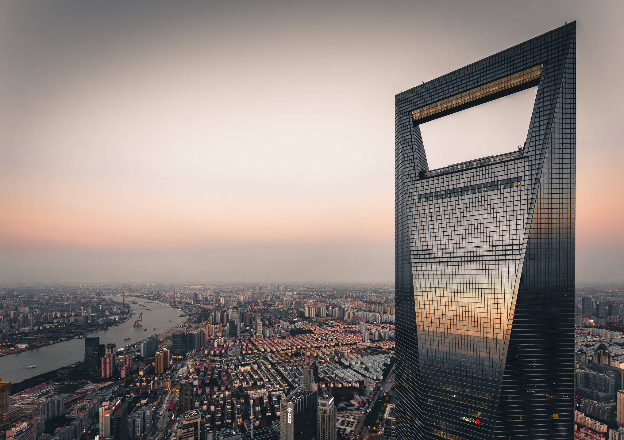 Shanghai 桌布 壁紙 — 一座高聳的摩天大樓帶有獨特的方形鏤空，在日落時分主宰著廣闊的城市景觀，其玻璃外牆在河流和密集的城市網格上反射出溫暖的色調。