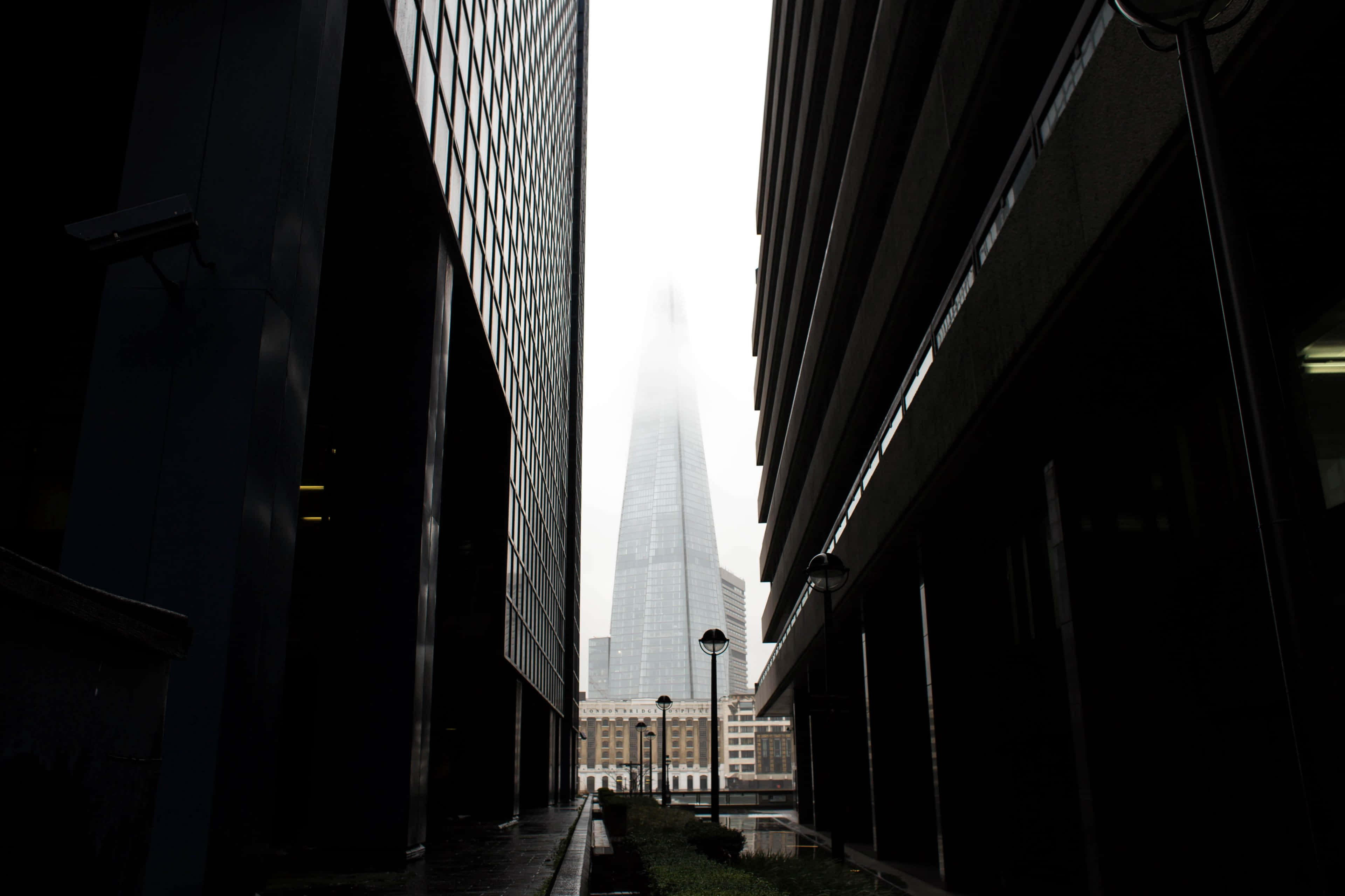 Buildings 桌布 壁紙 — 一座高聳的摩天大樓穿過霧濛濛的天空，在狹窄的城市小巷中被深色現代建築框住。