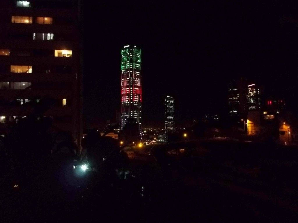Bogota 桌布 壁紙 — 一座高聳的摩天大樓被綠色、紅色和白色的燈光照亮，在繁華城市景觀的黑暗夜空中脫穎而出。