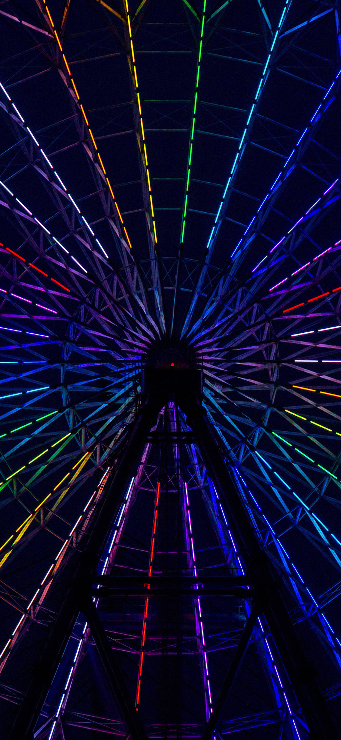 Neon Light 桌布 壁紙 — 一座高聳的摩天輪在夜晚散發出彩虹色調的鮮豔霓虹燈光，映襯著漆黑的天空。