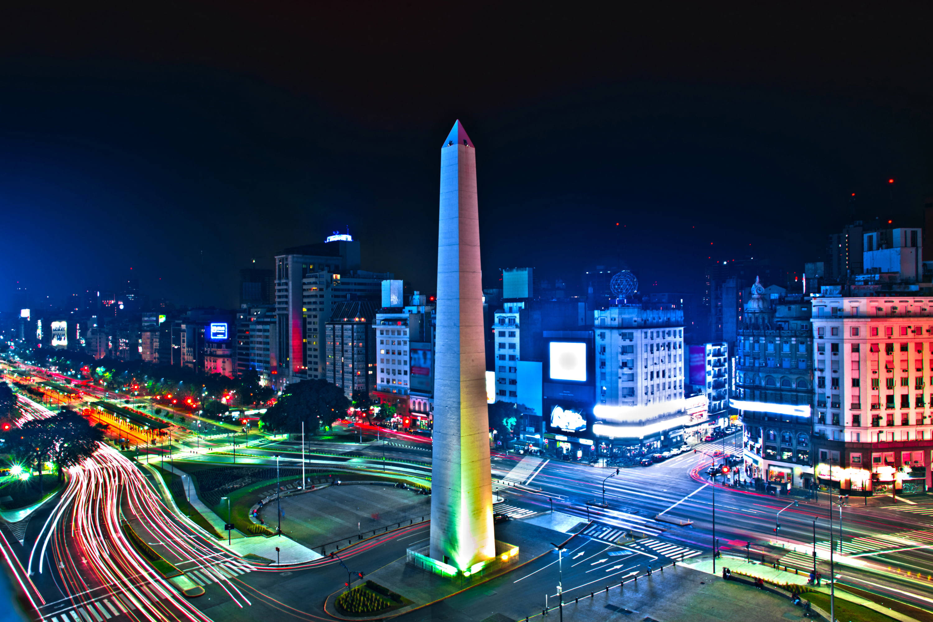 Argentina 桌布 壁紙 — 一座高聳的方尖碑在夜晚閃爍著彩虹光芒，周圍是繁華的城市景觀和車流燈軌。
