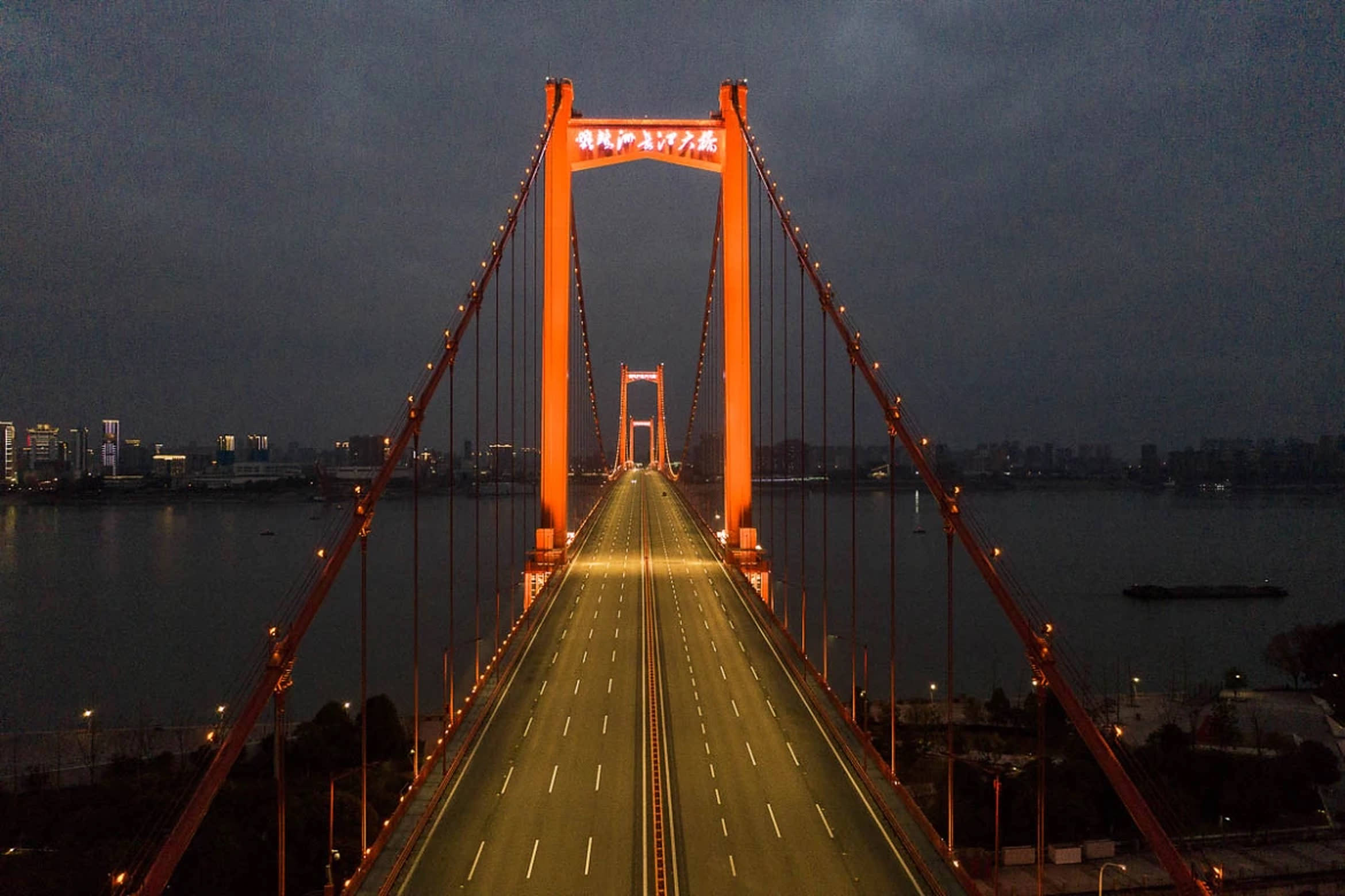 Wuhan 桌布 壁紙 — 一座高聳的橙色吊橋在暮色中發光，空蕩的車道延伸向遠方的城市天際線。