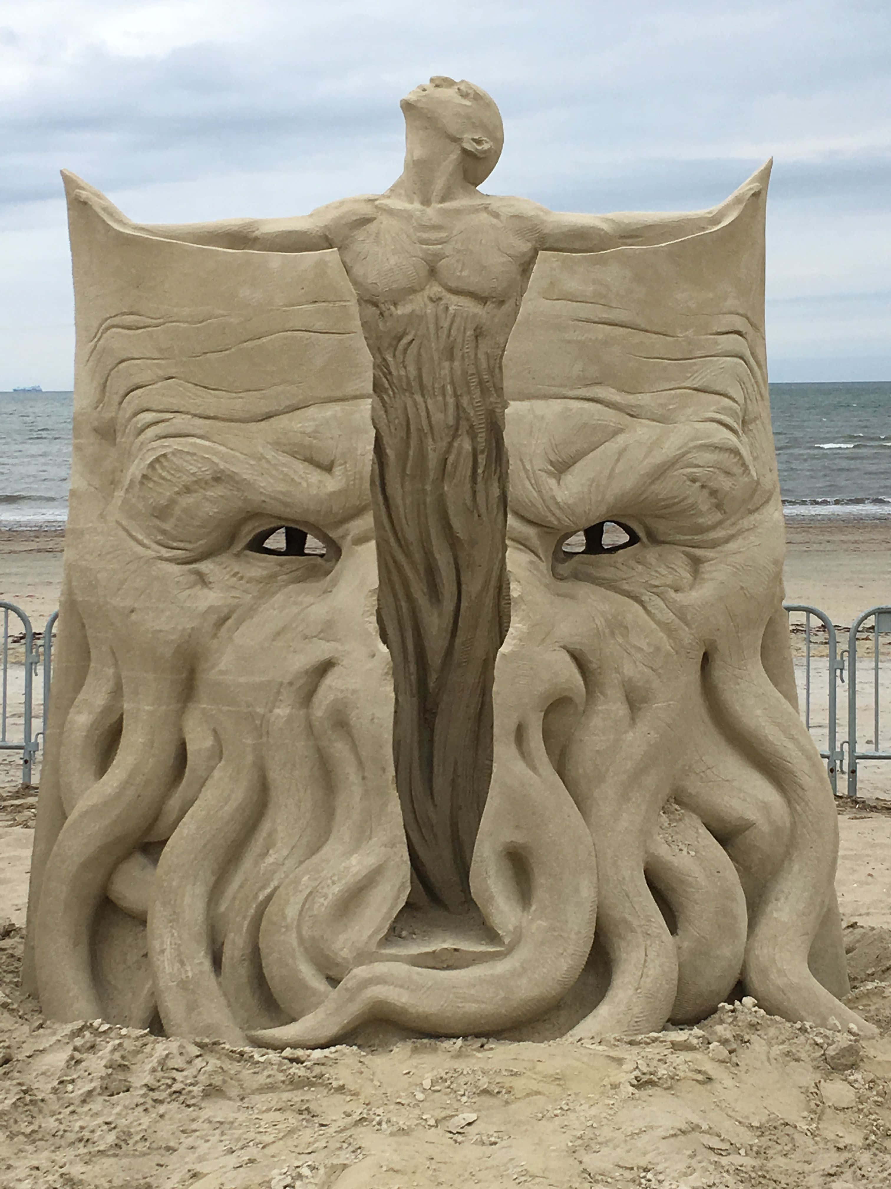 Sand Art 桌布 壁紙 — 一座高聳的沙雕描繪了一個肌肉發達的男人站在沙灘上兩個巨型章魚臉之間，眼神銳利。