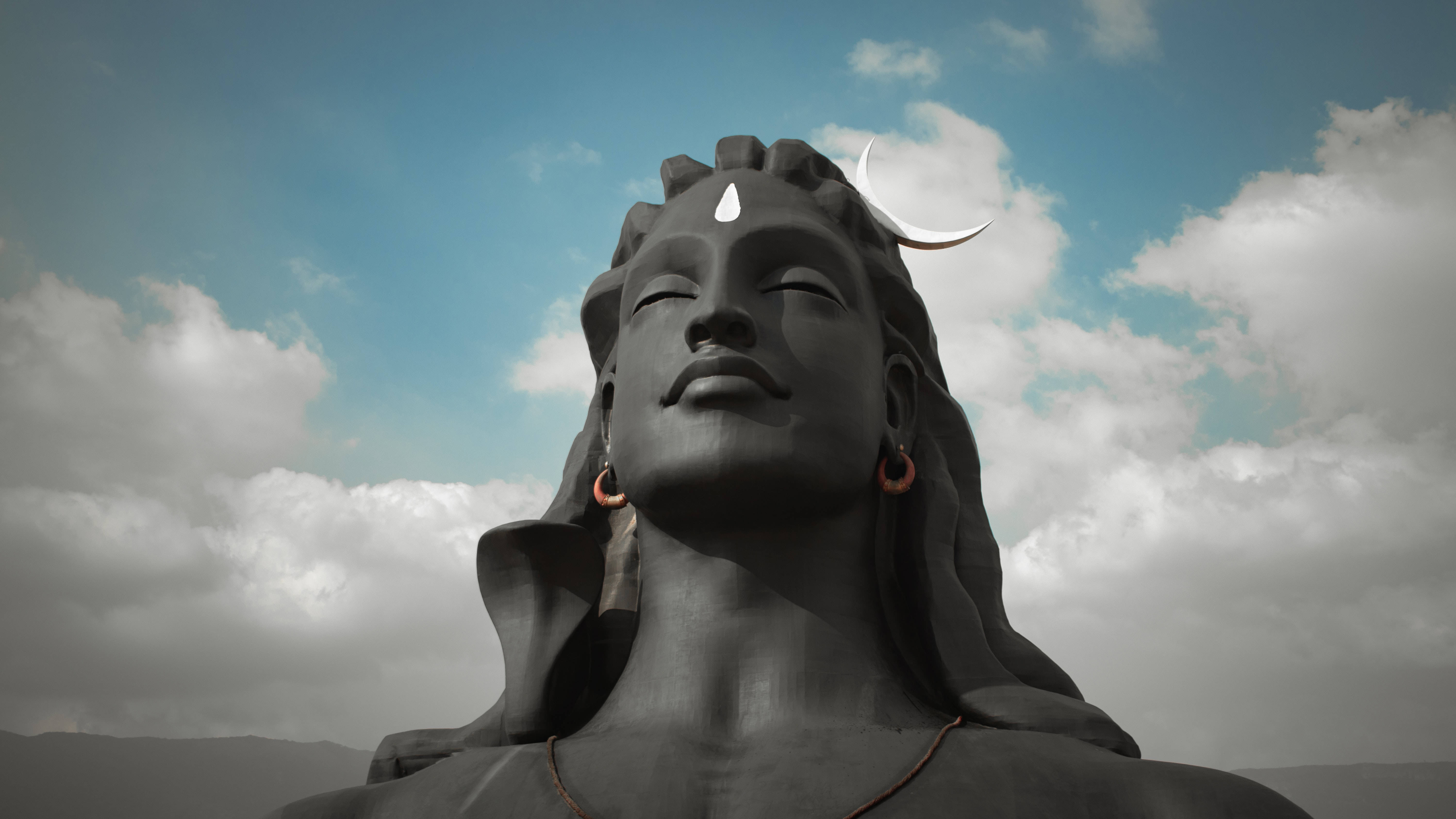 adiyogi shiva 桌布 壁紙 — 一座高聳的深色濕婆石像，頭戴新月冠冕，額有第三隻眼，在佈滿雲朵的藍天下顯得寧靜祥和。