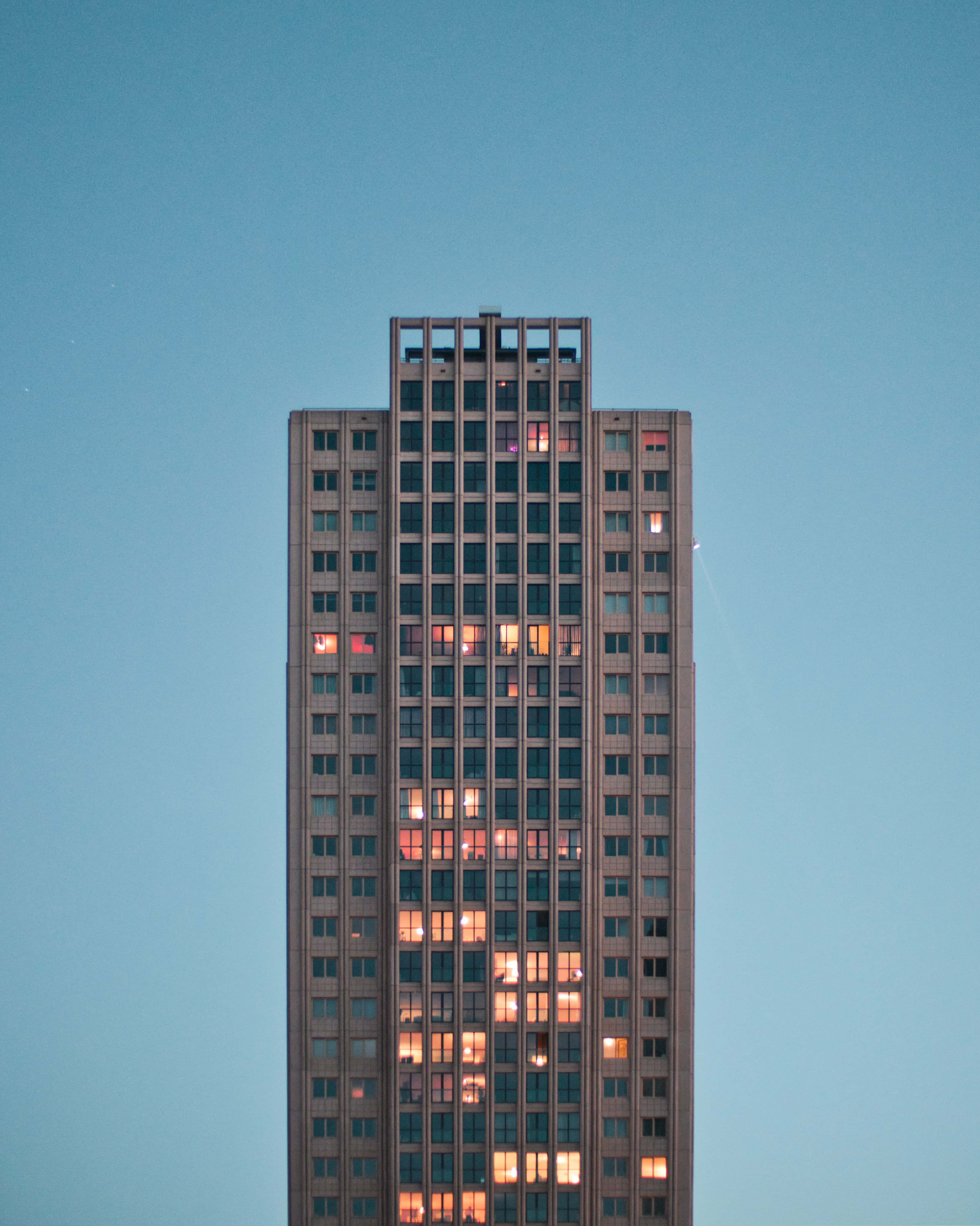 Building 桌布 壁紙 — 一座高聳的混凝土公寓大樓矗立在清澈的暮色天空下，散落的窗戶透出溫暖的燈光。
