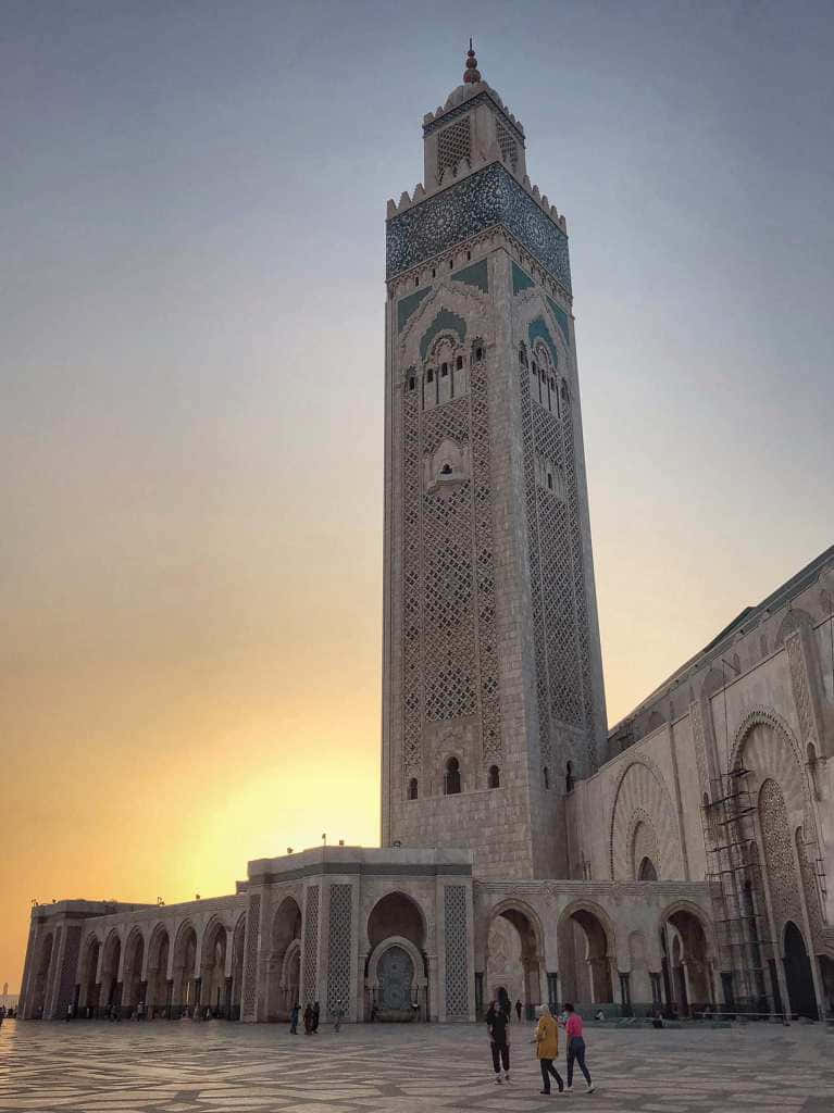 Mosque 桌布 壁紙 — 一座高聳的清真寺宣禮塔，帶有精緻的瓷磚工藝，矗立在溫暖的日落天空下，人們在其瓷磚庭院中漫步。
