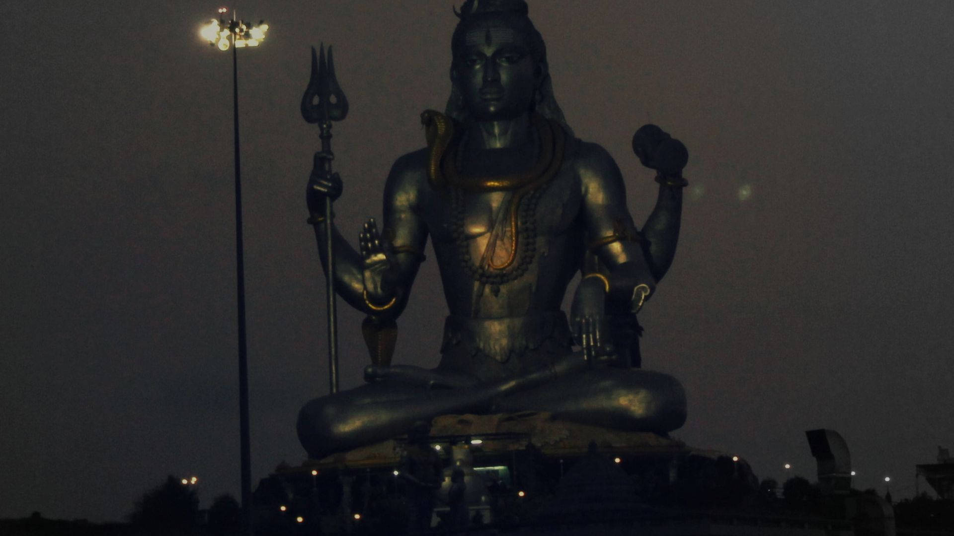 shiva dark 桌布 壁紙 — 一座高聳的濕婆神青銅像在夜空下冥想，附近街燈閃爍。