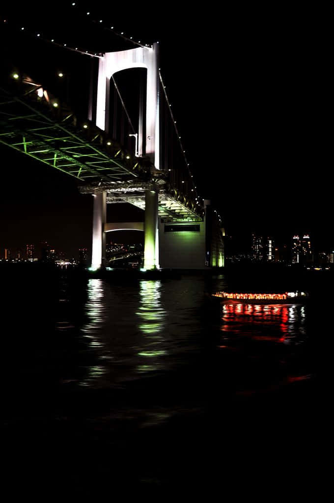 Rainbow Bridge 桌布 壁紙 — 一座高聳的照明橋樑在夜間倒映在黑暗的水面上，附近有一艘發光的船。