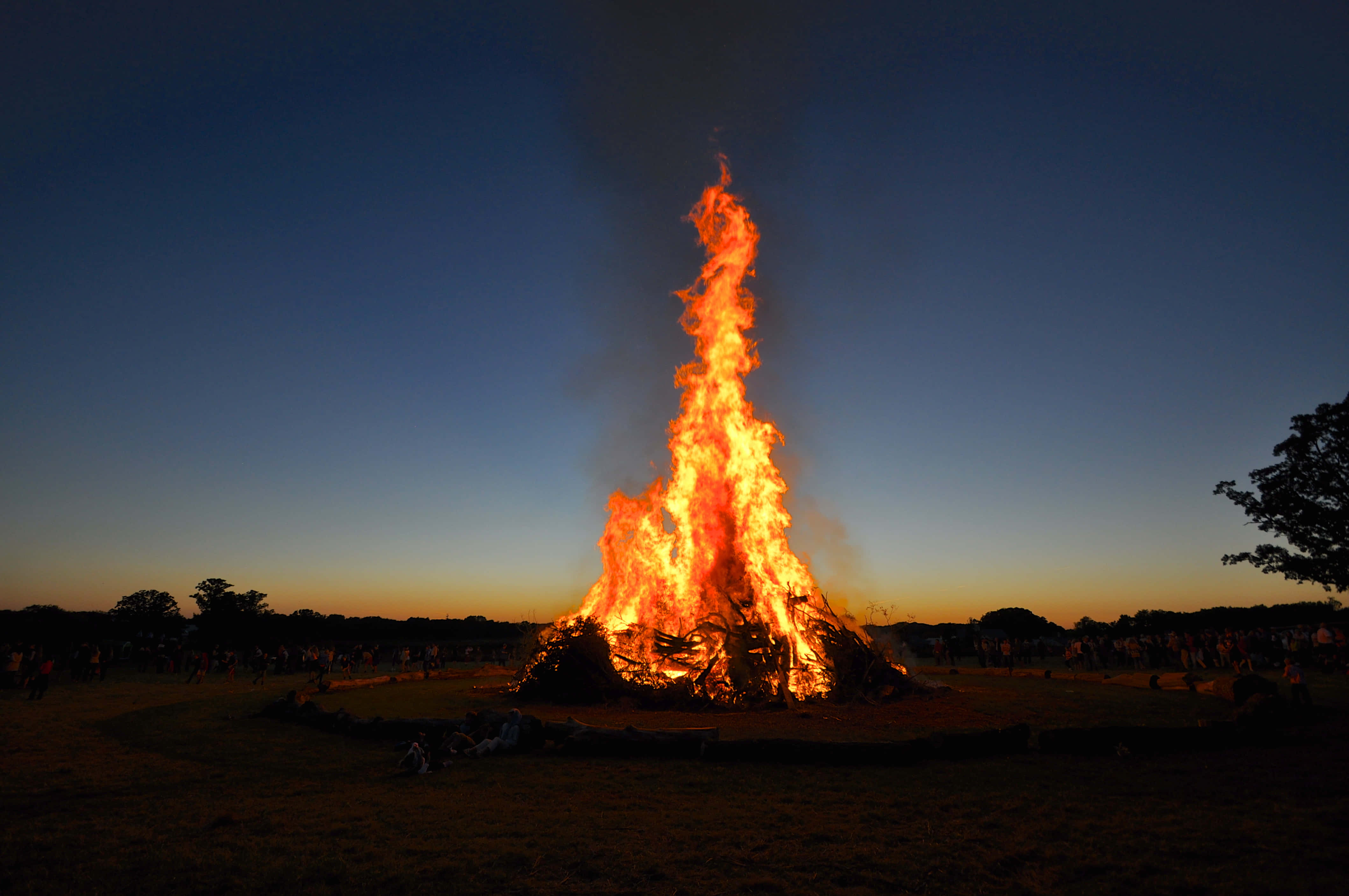 Fall Bonfire 桌布 壁紙 — 一座高聳的營火在黃昏時分熊熊燃燒，將熾熱的橙色光芒投射在草地上，觀眾的身影呈現剪影。