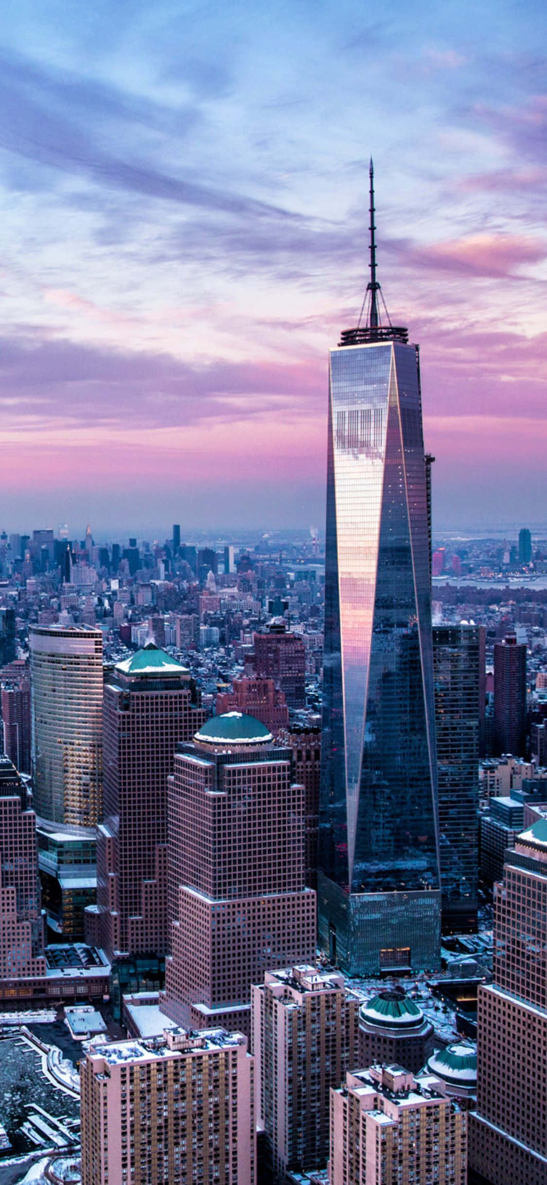 iPhone X New York 桌布 壁紙 — 一座高聳的玻璃帷幕摩天大樓，在充滿活力的城市天際線中反射出粉紅色和紫色的日落色調。