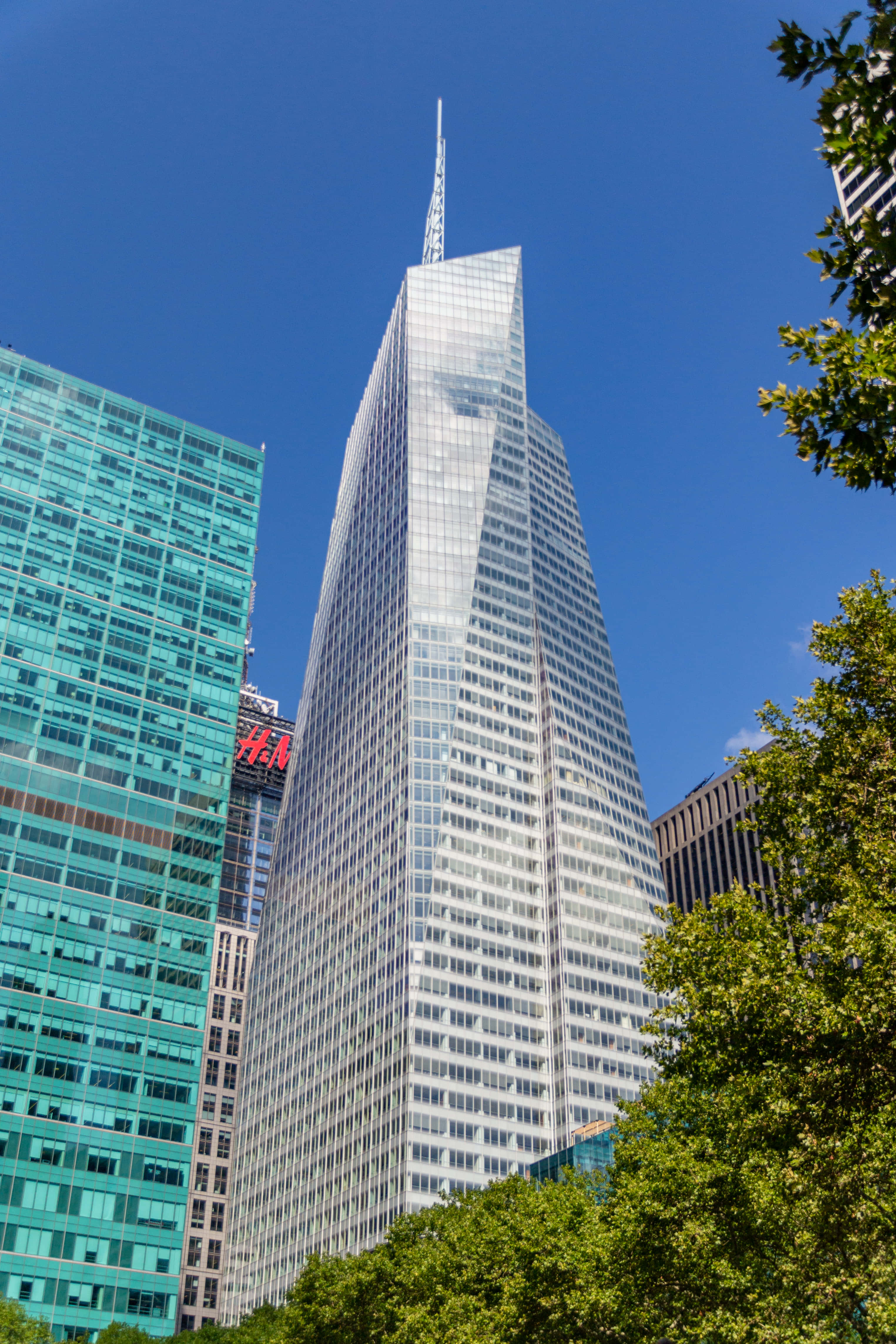 Bank Of America 桌布 壁紙 — 一座高聳的玻璃摩天大樓，帶有尖銳的尖頂，刺破清澈的藍天，周圍環繞著茂密的綠樹，兩側是其他現代建築。