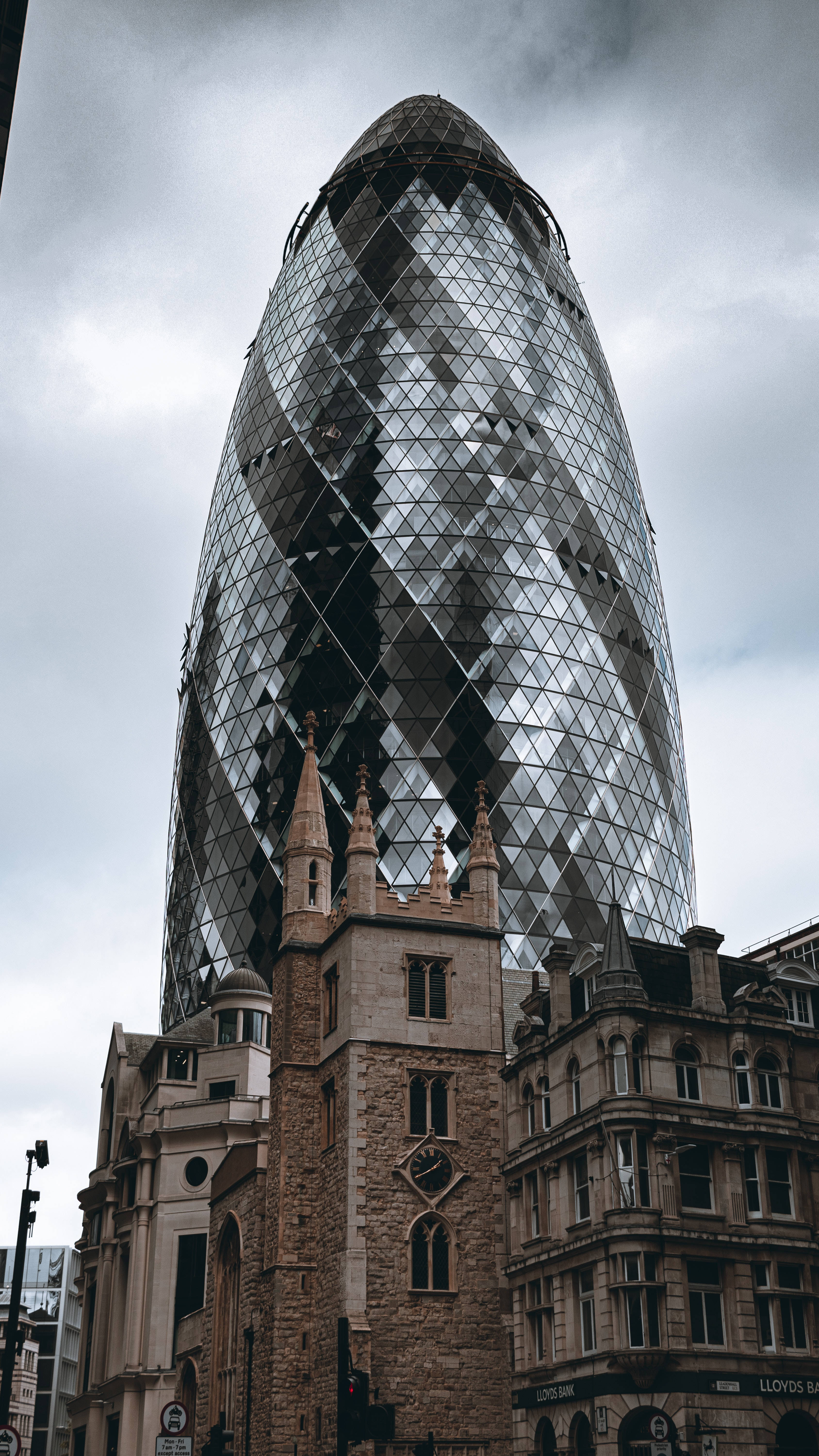 Buildings 桌布 壁紙 — 一座高聳的玻璃摩天大樓，帶有獨特的彎曲鑽石圖案，在陰天天空下聳立於歷史悠久的石砌建築之上。