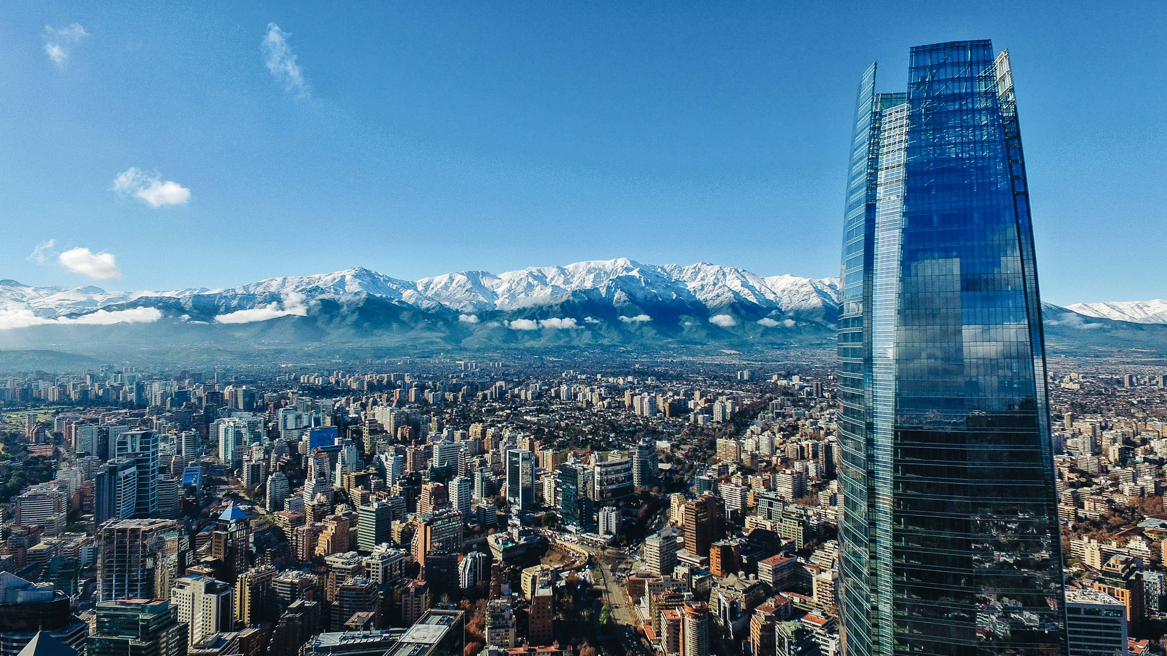 Chile 桌布 壁紙 — 一座高聳的玻璃摩天大樓主宰著廣闊的城市景觀，背後是白雪覆蓋的山脈，在清澈的藍天下崛起。